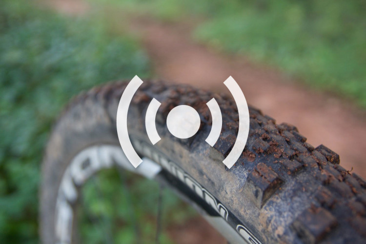 Close-up of a mountain bike tire with visible tread patterns and some mud on it, set against a blurred green background. An abstract icon, symbolizing connectivity, is superimposed in the center of the image.