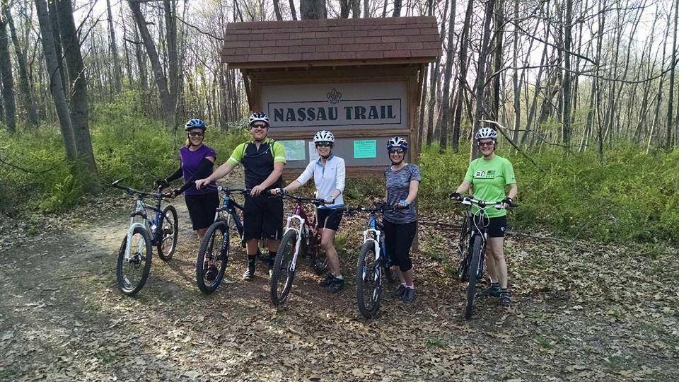 Five mountain bikers pose with their bikes in front of a sign for the Nassau Trail, surrounded by a lush forest in early spring. Each person is wearing a helmet and cycling attire, smiling and ready for their adventure.