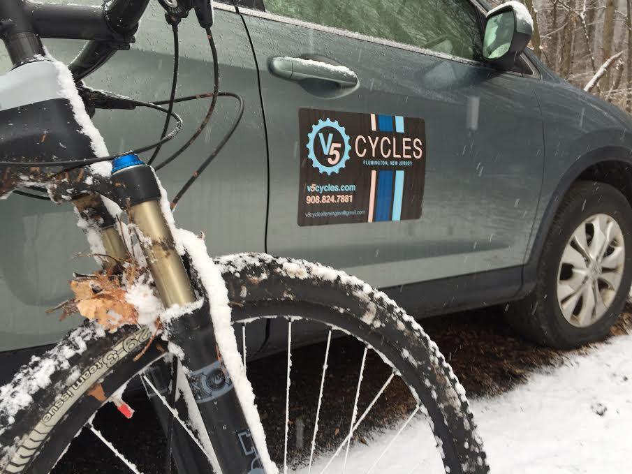 A close-up view of a bicycle parked next to a car in a snowy setting. The car features a logo and contact information for 'V5 Cycles' located in Flemington, New Jersey. The bicycle's front wheel is partially covered in snow, and there are remnants of leaves stuck to the frame. Snowflakes are visibly falling in the scene.
