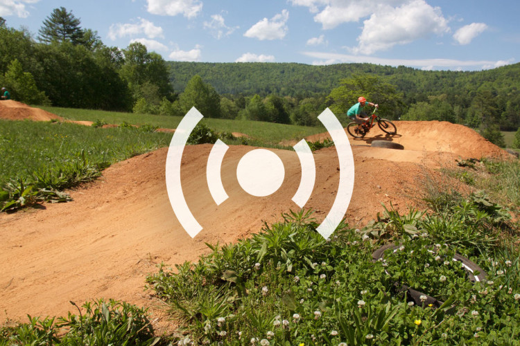 A cyclist performing a jump on a dirt bike track surrounded by green grass and trees under a blue sky with fluffy clouds.