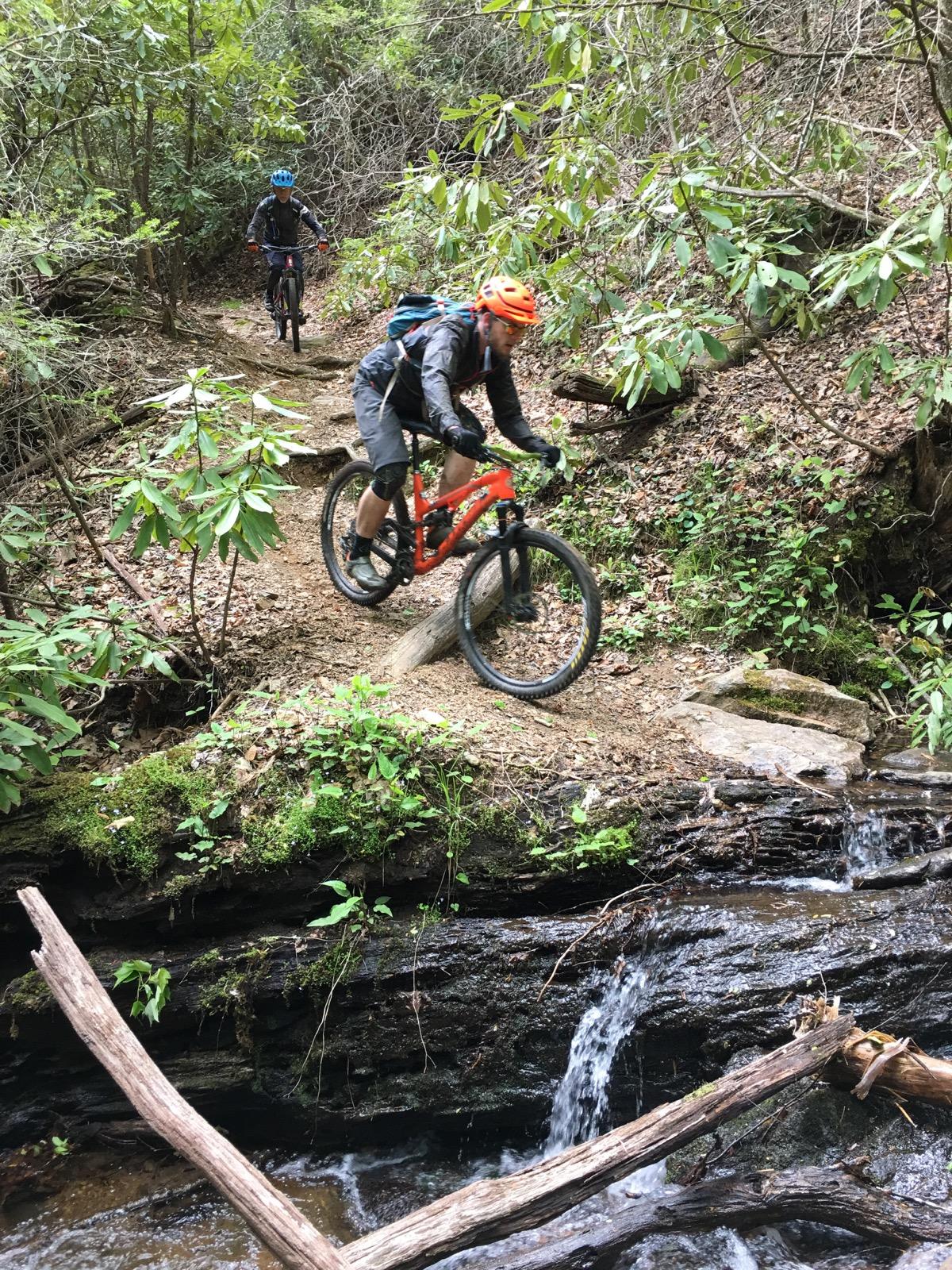 Two mountain bikers navigate a narrow trail in a dense forest. One cyclist, wearing an orange helmet and a backpack, is riding over a rocky area near a small stream. The second biker, in the background, is following the same path. Lush green foliage surrounds the trail, creating a vibrant natural setting. Slate Rock (102) mountain bike trail.