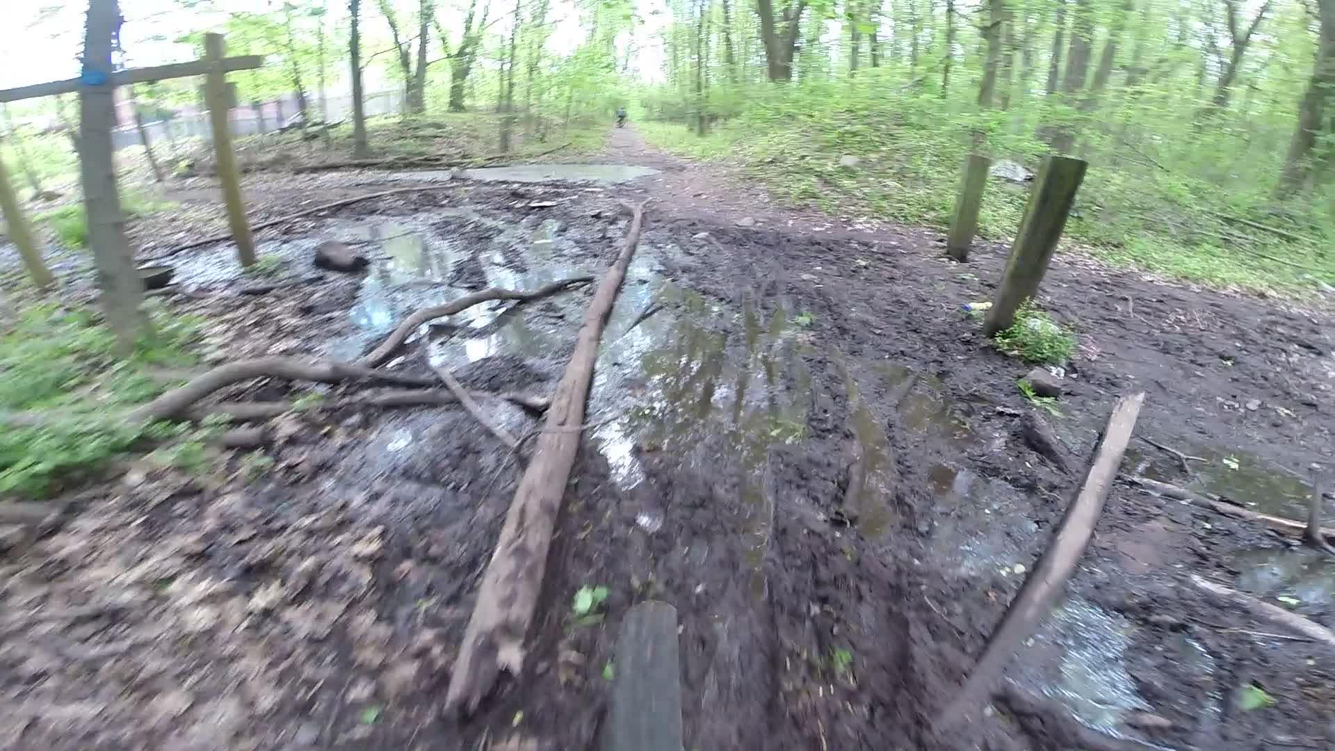 A muddy forest path with puddles and fallen branches, surrounded by lush green trees. The trail leads into the distance, with a hint of another person walking further along the path. Richmond Avenue and Forest Hill road mountain bike trail.