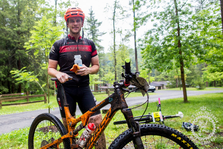 A muddy mountain biker stands beside an orange bike in a forested area. He is wearing a black cycling jersey with red accents, a helmet, and has dirt on his face. In one hand, he holds a bottle of drink and a snack, while the other hand grips the bike. Lush green trees and a gravel path are visible in the background.