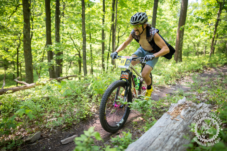 A mountain biker in a tank top and denim shorts rides a trail through a lush green forest. The cyclist wears a helmet and sunglasses, and is focused on navigating the rocky terrain. Sunlight filters through the trees, highlighting the vibrant greenery around the path. A log lies on the trail as the biker approaches. A race number is visible on the front of the bike.