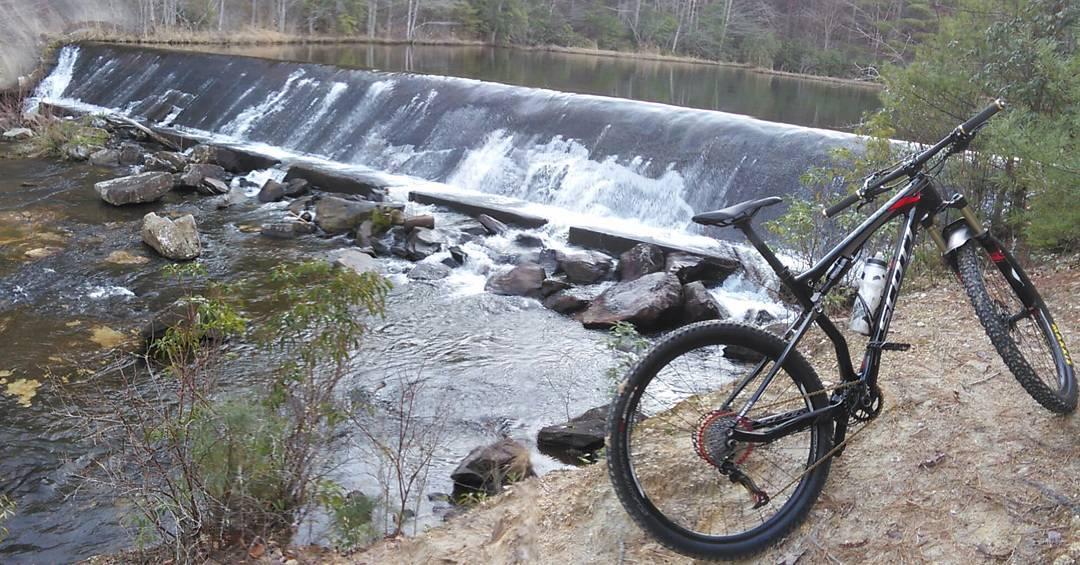 Scott Spark 910: A mountain bike rests beside a flowing river with a waterfall over a dam in the background. The scene is set in a wooded area, showcasing rocks and greenery along the water's edge on a cloudy day.