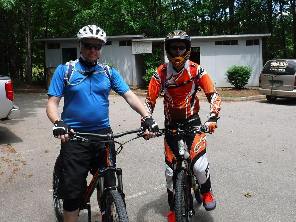 Two mountain bikers pose for a photo in a wooded area. One rider is wearing a blue shirt and black shorts, while the other is dressed in bright orange and black protective gear. Both are wearing helmets and holding their bikes, with a bike wash facility visible in the background. The scene is set in a natural environment, indicating an outdoor biking activity. Chicopee Woods mountain bike trail.