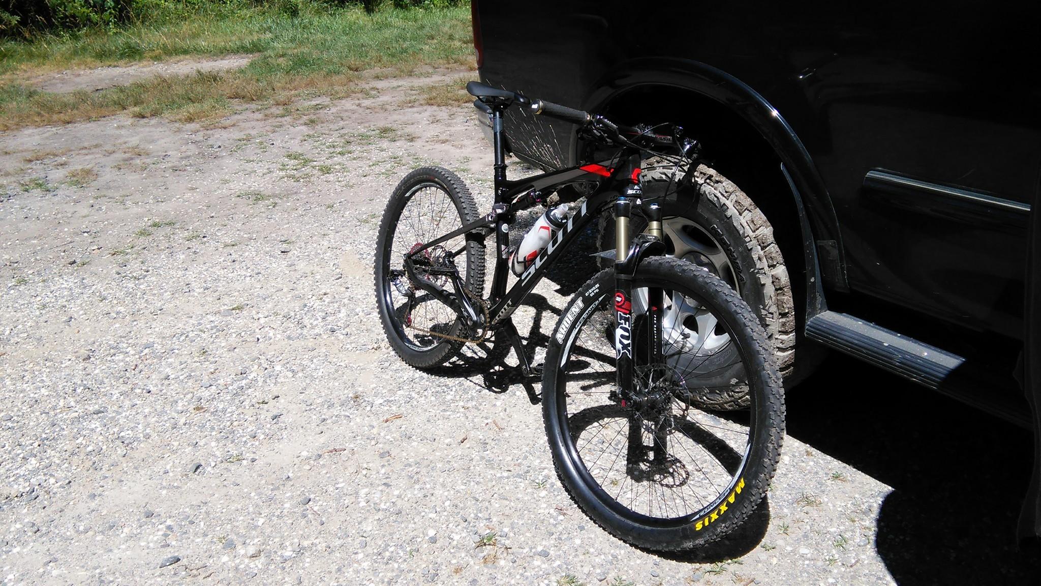 Scott Spark 910: A black and red mountain bike with wide tires is leaning against the rear of a dark vehicle on a gravel surface. The bike features a water bottle holder and is positioned in a natural outdoor setting with grass and trees in the background.