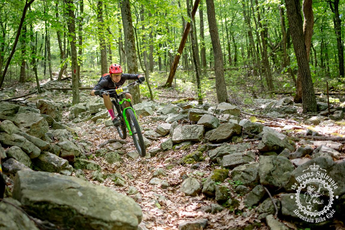 A mountain biker navigating a rocky trail in a lush green forest. The rider is wearing a bright helmet and colorful socks, leaning forward on a vibrant green bike as they skillfully maneuver over uneven terrain. Sunlight filters through the trees, creating a vibrant outdoor atmosphere. Wildcat Gap mountain bike trail.