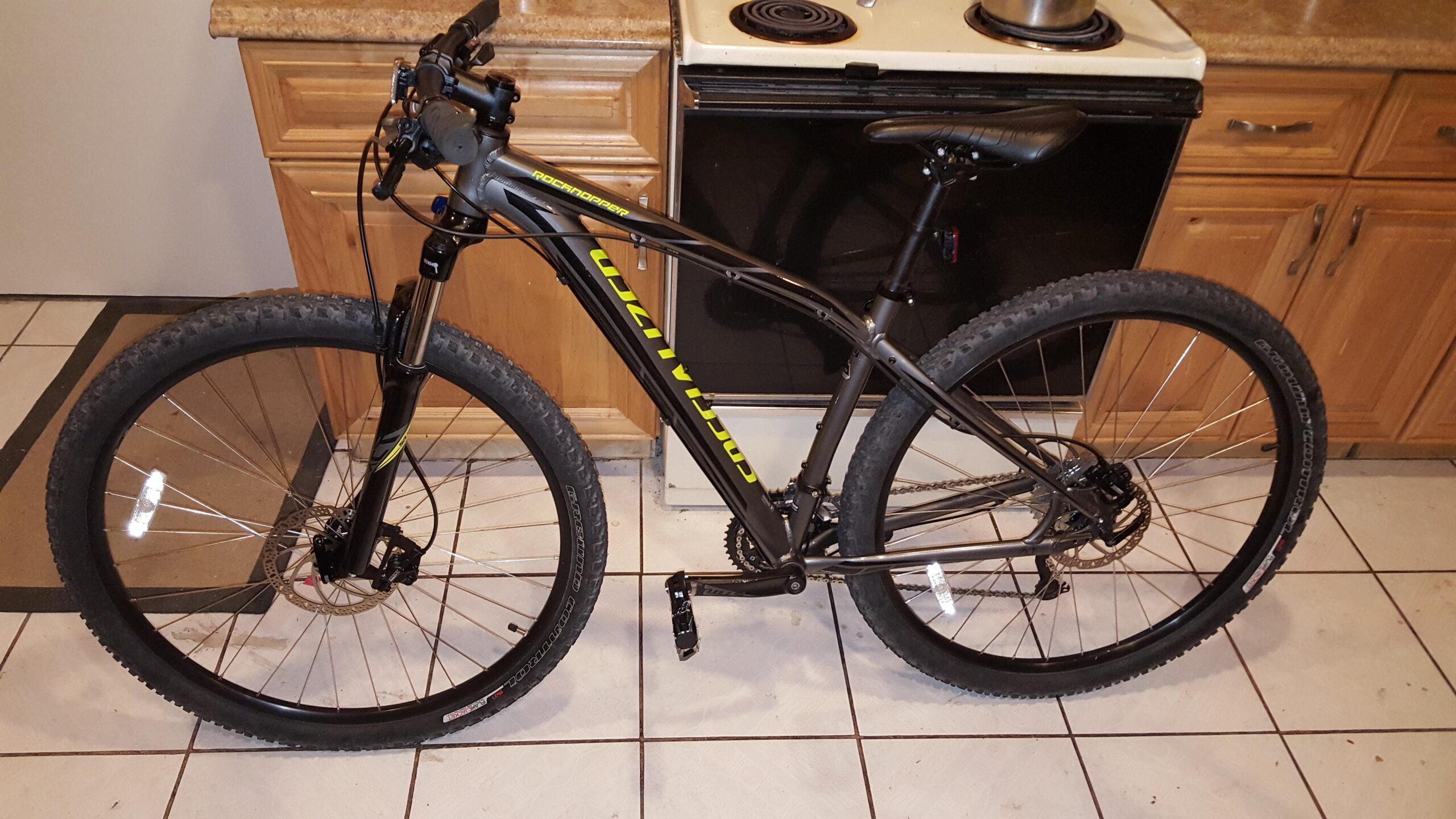 Specialized Rockhopper 29: A black mountain bike with yellow accents, positioned indoors on a tiled floor. The bike features large tires, a front suspension fork, and a visible chain and gears. In the background, a kitchen area is partially visible, including wooden cabinetry and a stove.