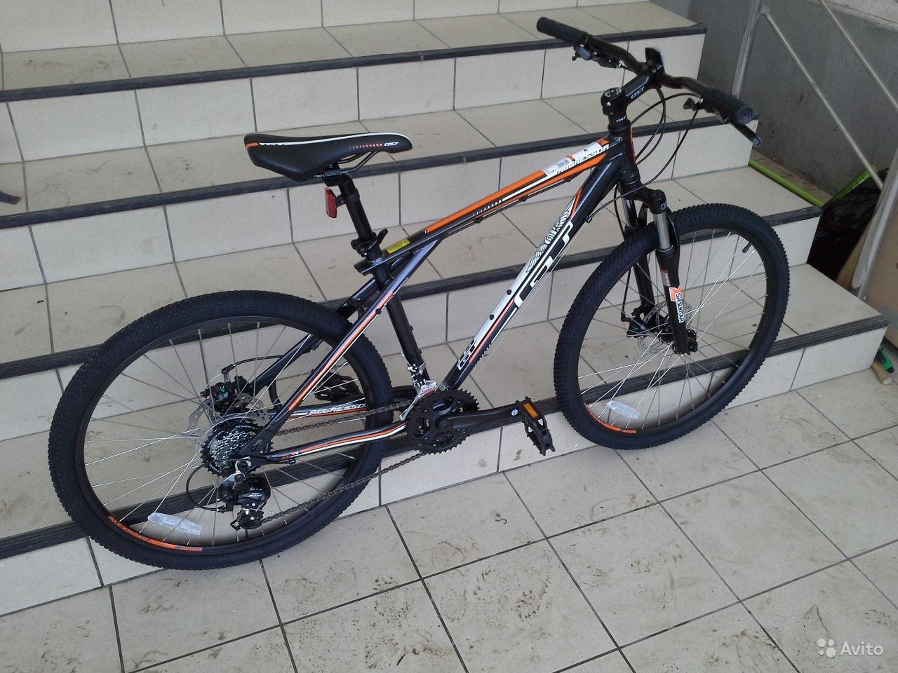 A black and orange mountain bike positioned on a tiled floor near a set of stairs. The bike features a sturdy frame, thick tires, and a comfortable seat, showcasing its off-road capabilities.