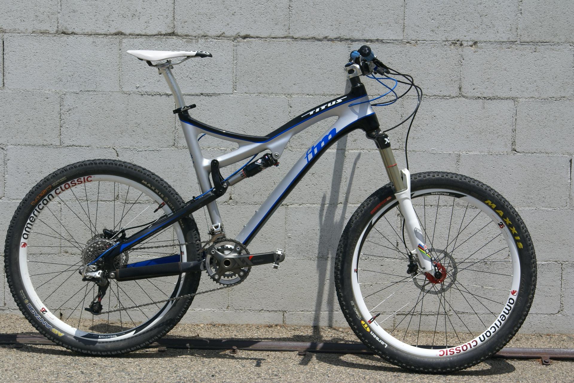 Titus FTM Carbon: Alt text: A blue and silver mountain bike leaning against a gray concrete wall. The bike features a lightweight frame, rugged tires, and front suspension, showcasing its design for off-road cycling.