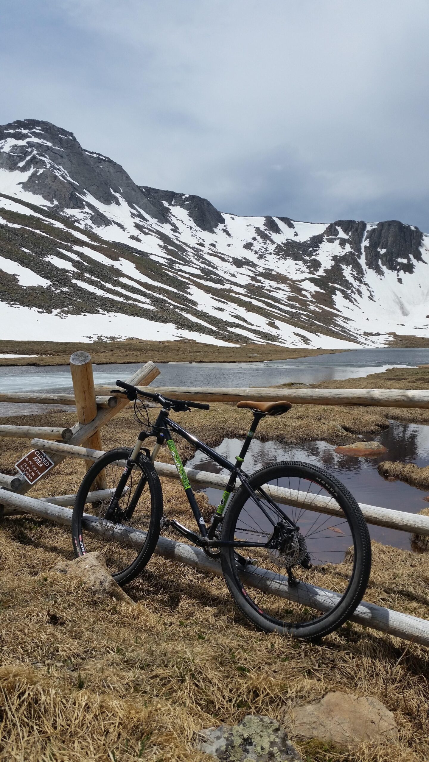 Salsa El Mariachi 3: A mountain bike leans against a wooden fence near a small body of water, with snow-capped mountains in the background. The area features patchy grass and rocky terrain, suggesting a remote, natural setting. A "Closed Area" sign is visible nearby.