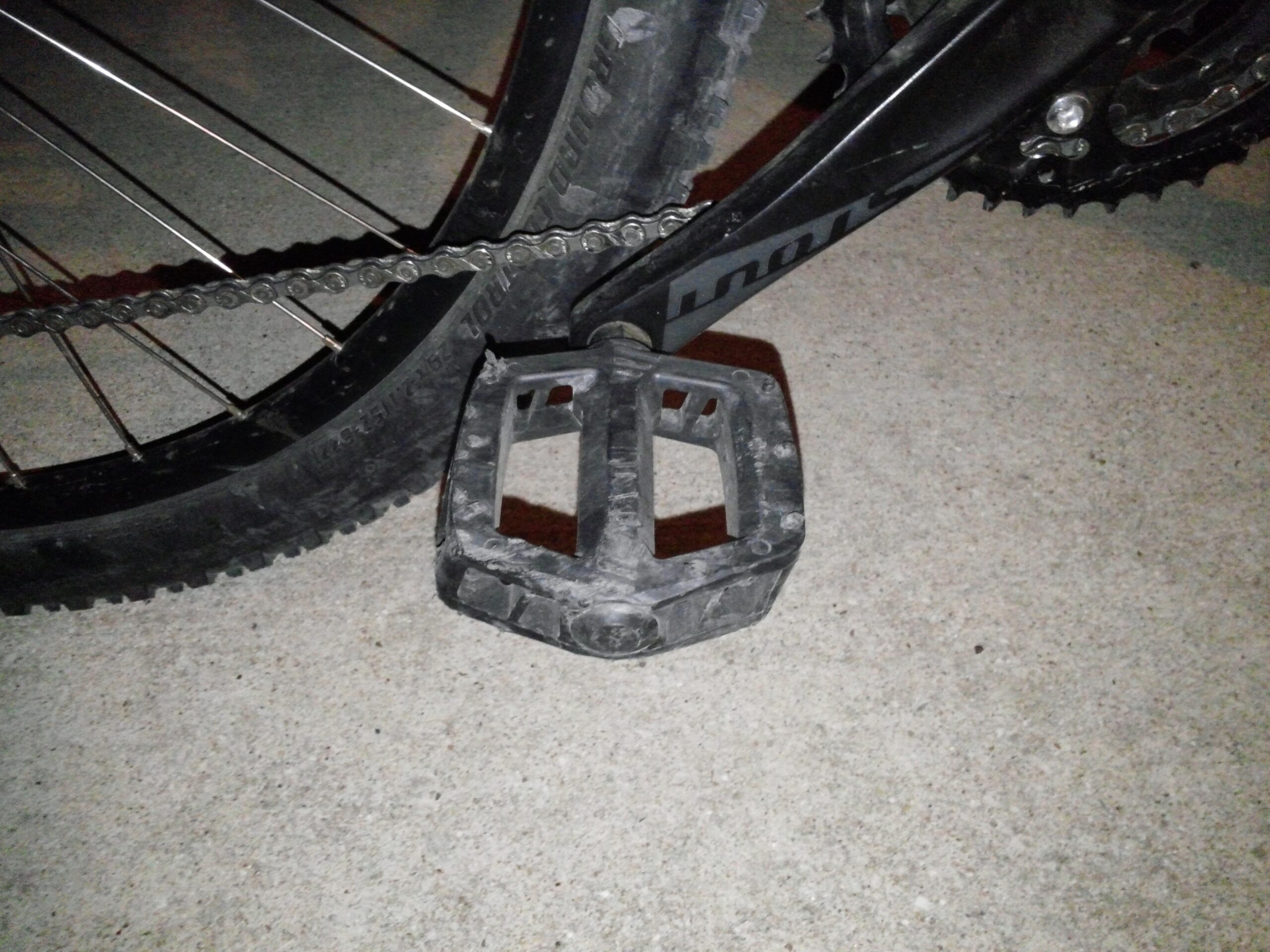 Specialized Rockhopper 29: Close-up of a bicycle pedal attached to a bike, with a chain and rear wheel partially visible. The image is taken on a concrete surface, showing the pedal's textured surface and the surrounding components.