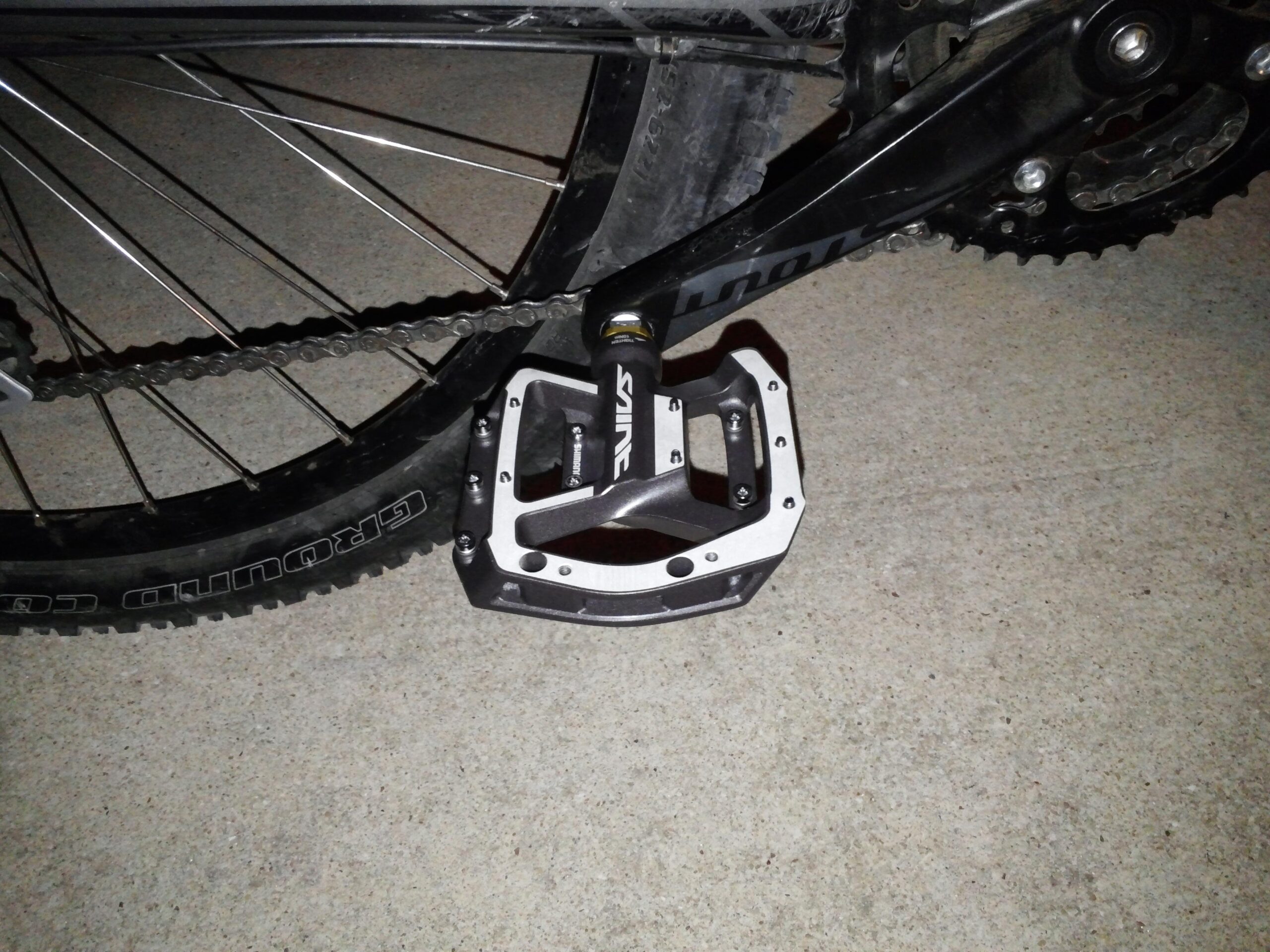 Specialized Rockhopper 29: Close-up of a bike pedal mounted on a bicycle crank, showing the pedal's metallic structure and distinct design. The background features the bike chain and rear wheel. The surface beneath is concrete.