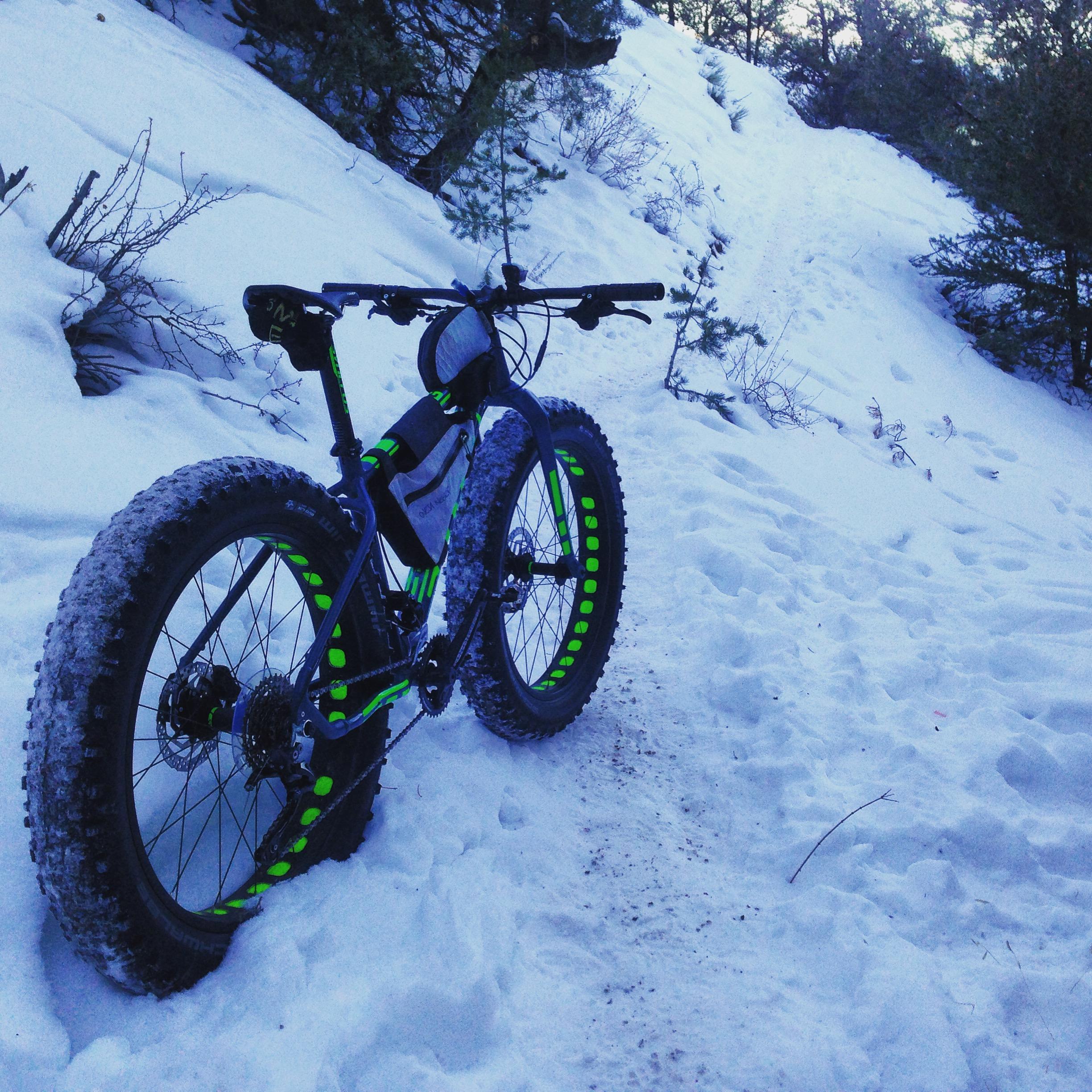 A fat bike with bright green accents is parked on a snowy trail surrounded by pine trees. The bike's large tires are covered in snow, and a narrow path leads up a slope in the background. North Backbone mountain bike trail.