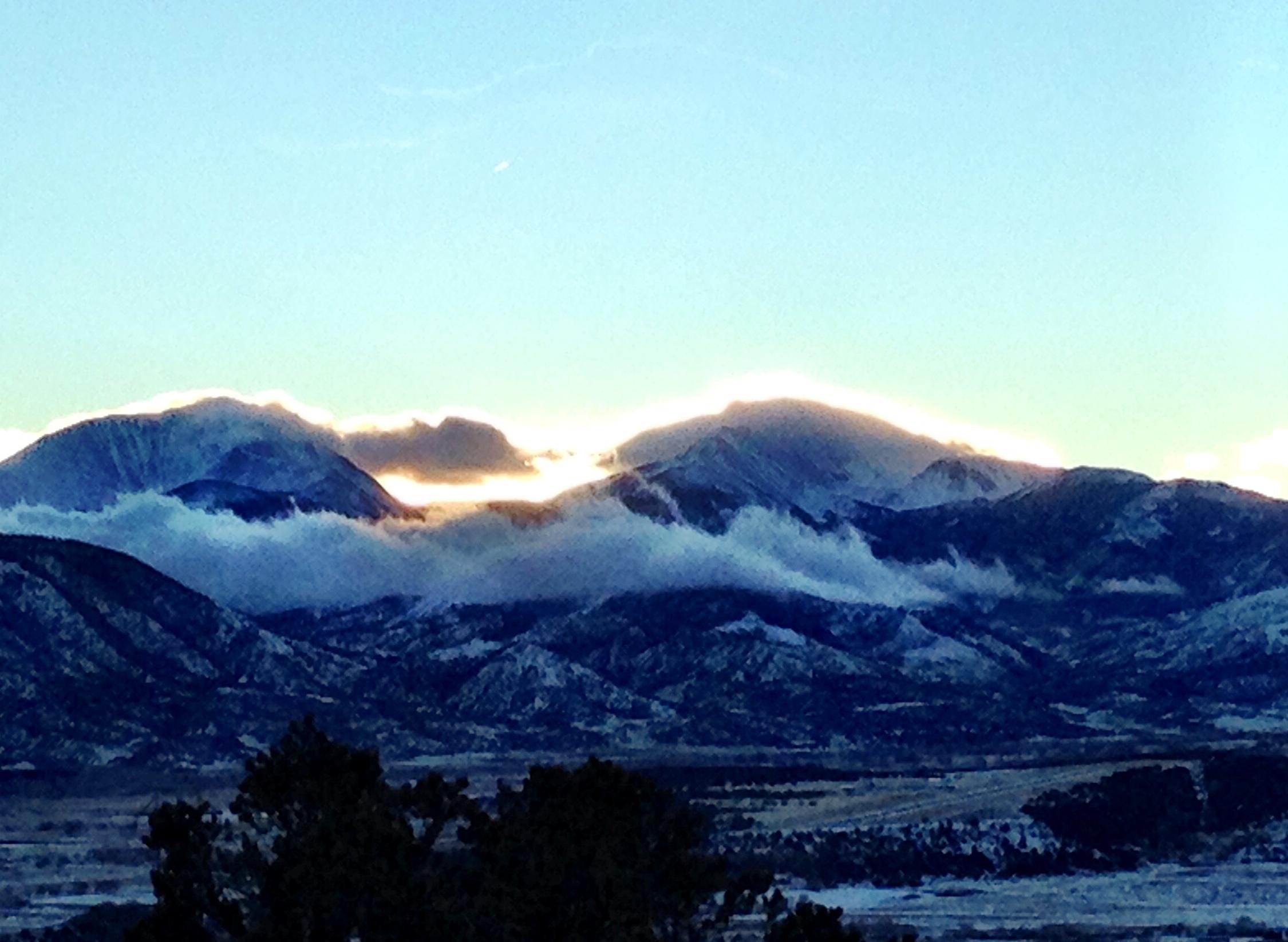A scenic view of snow-capped mountains at dawn, with soft clouds rolling through the valleys. The sky is painted in soft hues of blue and gold as the sun rises behind the peaks. A silhouette of dark trees sits in the foreground, providing depth to the landscape. North Backbone mountain bike trail.