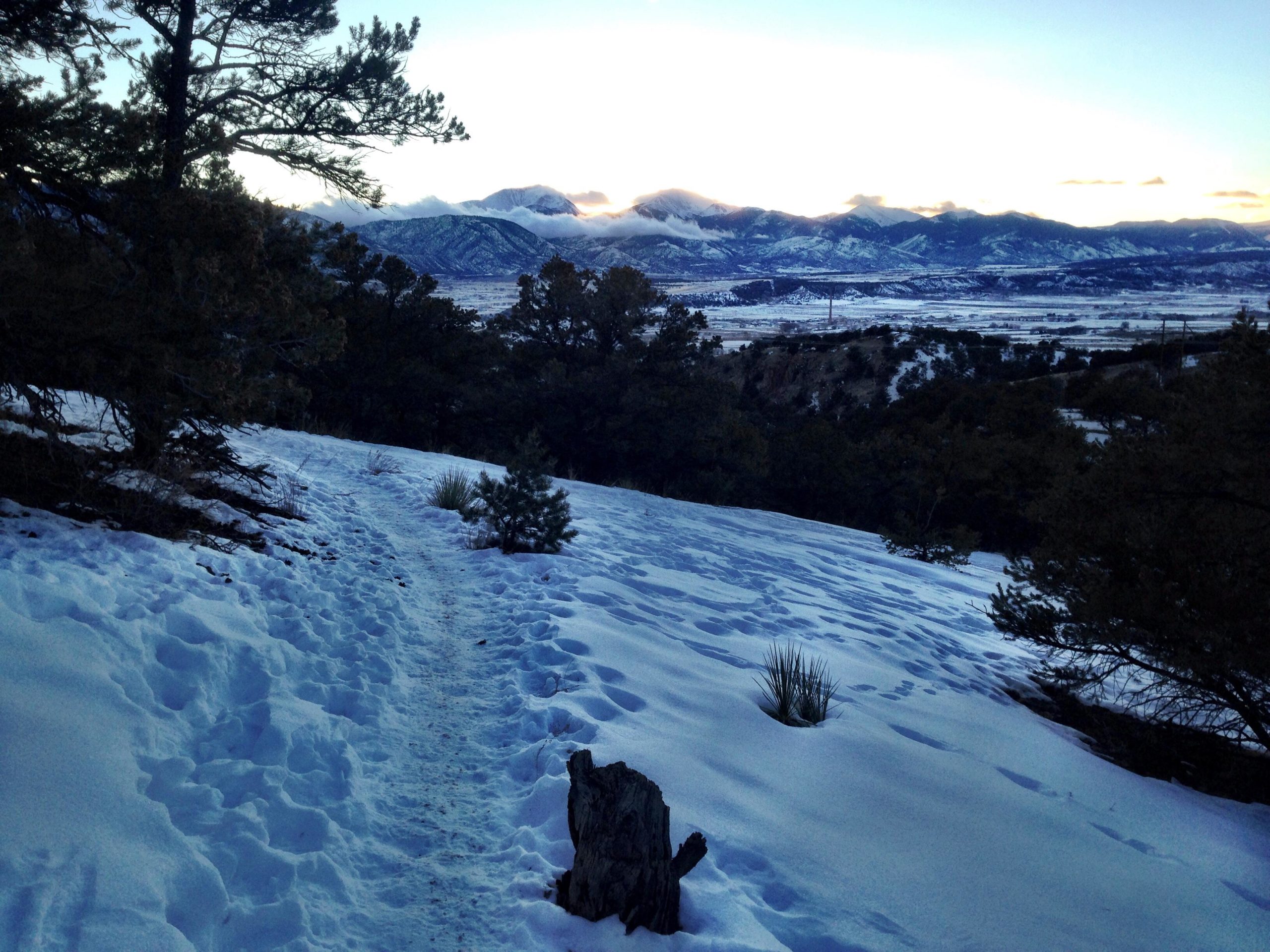 A snowy landscape with a winding dirt path leading through the snow, flanked by trees on either side. In the background, mountains are visible, their peaks dusted with snow under a partly cloudy sky. The scene captures a tranquil winter setting at dusk. North Backbone mountain bike trail.