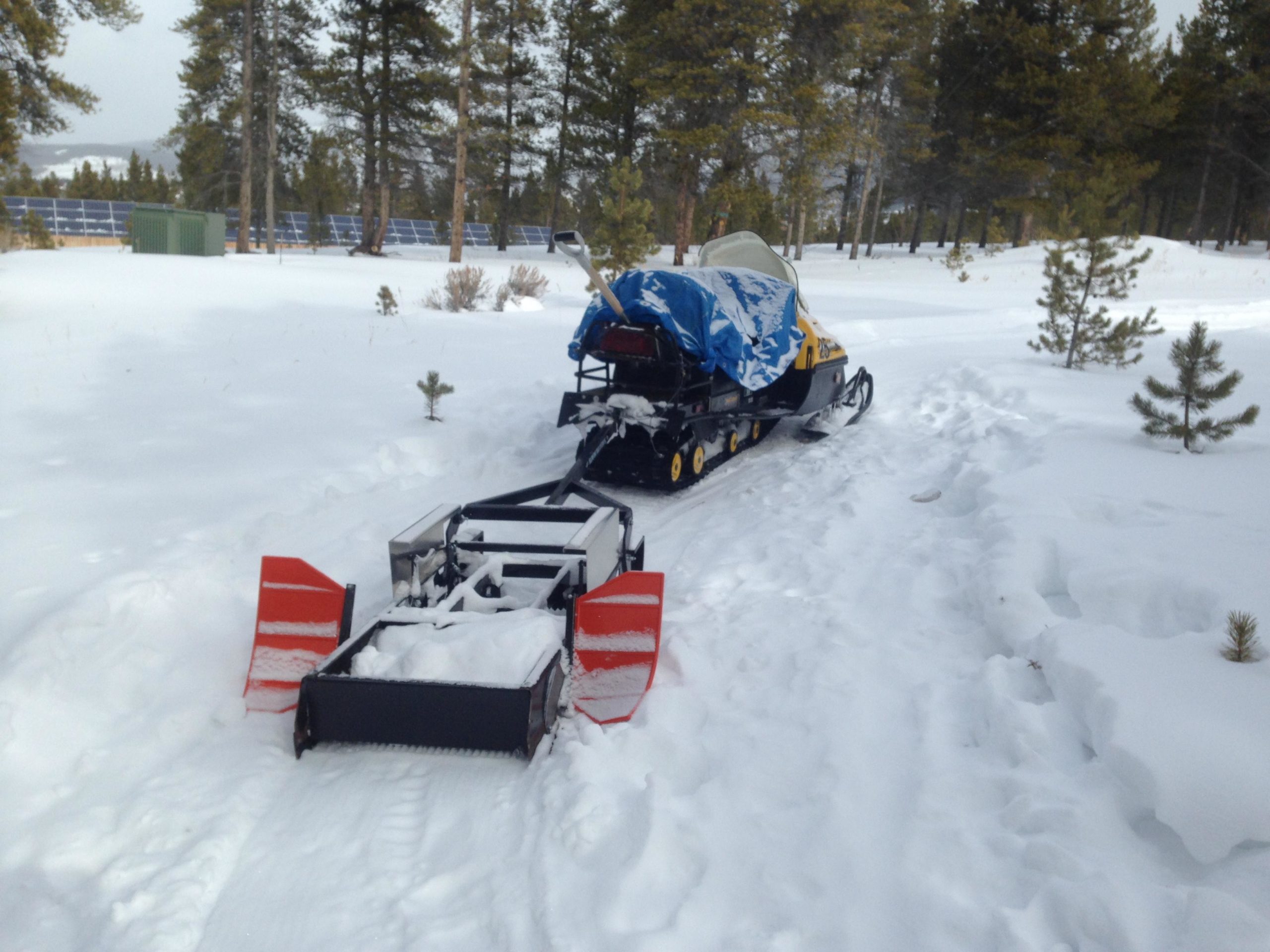 A snowmobile parked on a snowy path, equipped with a snow plow attachment, surrounded by pine trees in a winter landscape. In the background, there are solar panels and a small structure. The ground is covered in thick snow, indicating a cold environment. Colorado Mountain College Trails mountain bike trail.
