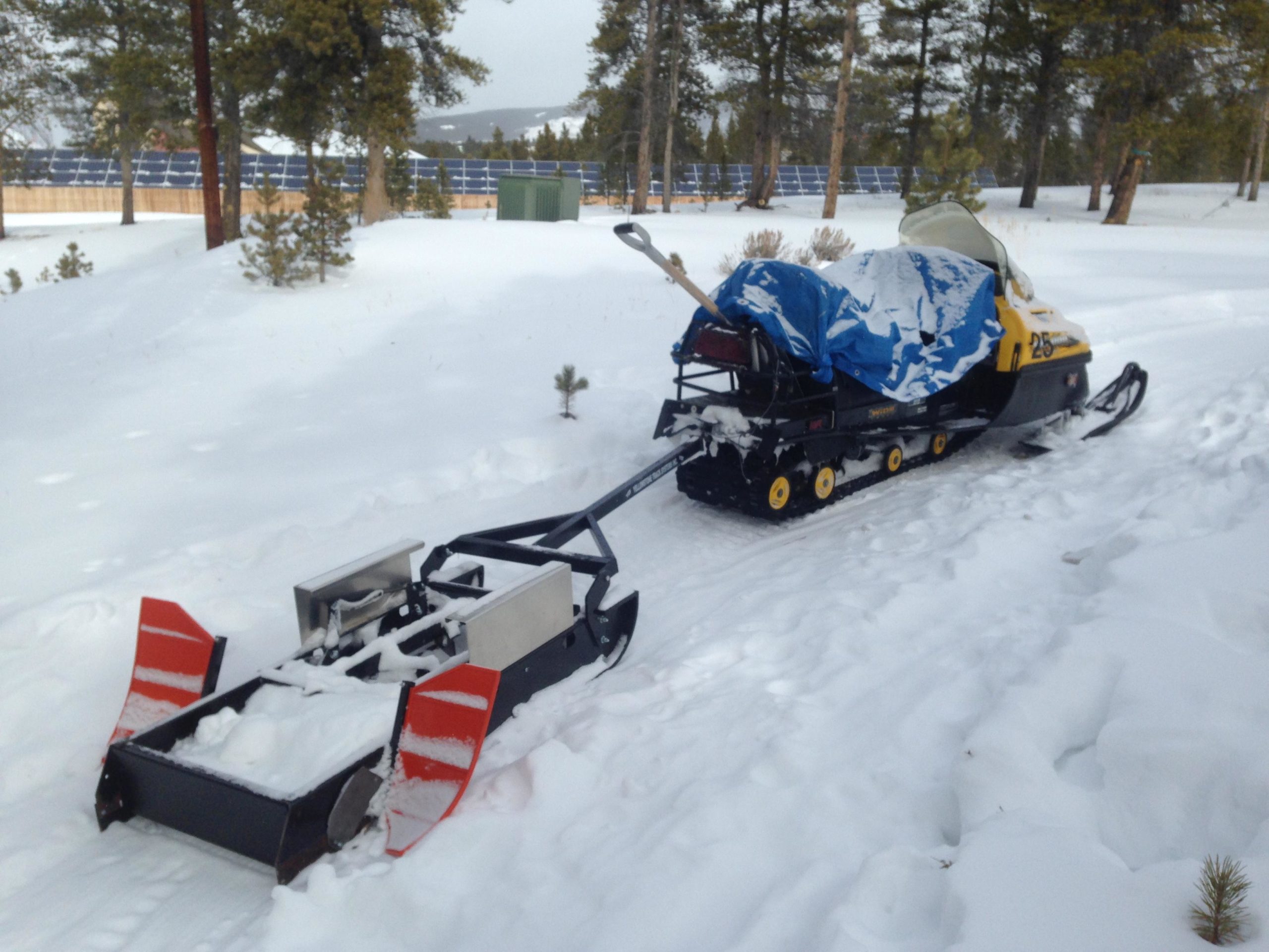 A snowmobile with a blue tarp covering it is parked in a snowy landscape, attached to a snow plow. The surrounding area features patches of snow and trees, with a background of solar panels and a building partially visible. Colorado Mountain College Trails mountain bike trail.