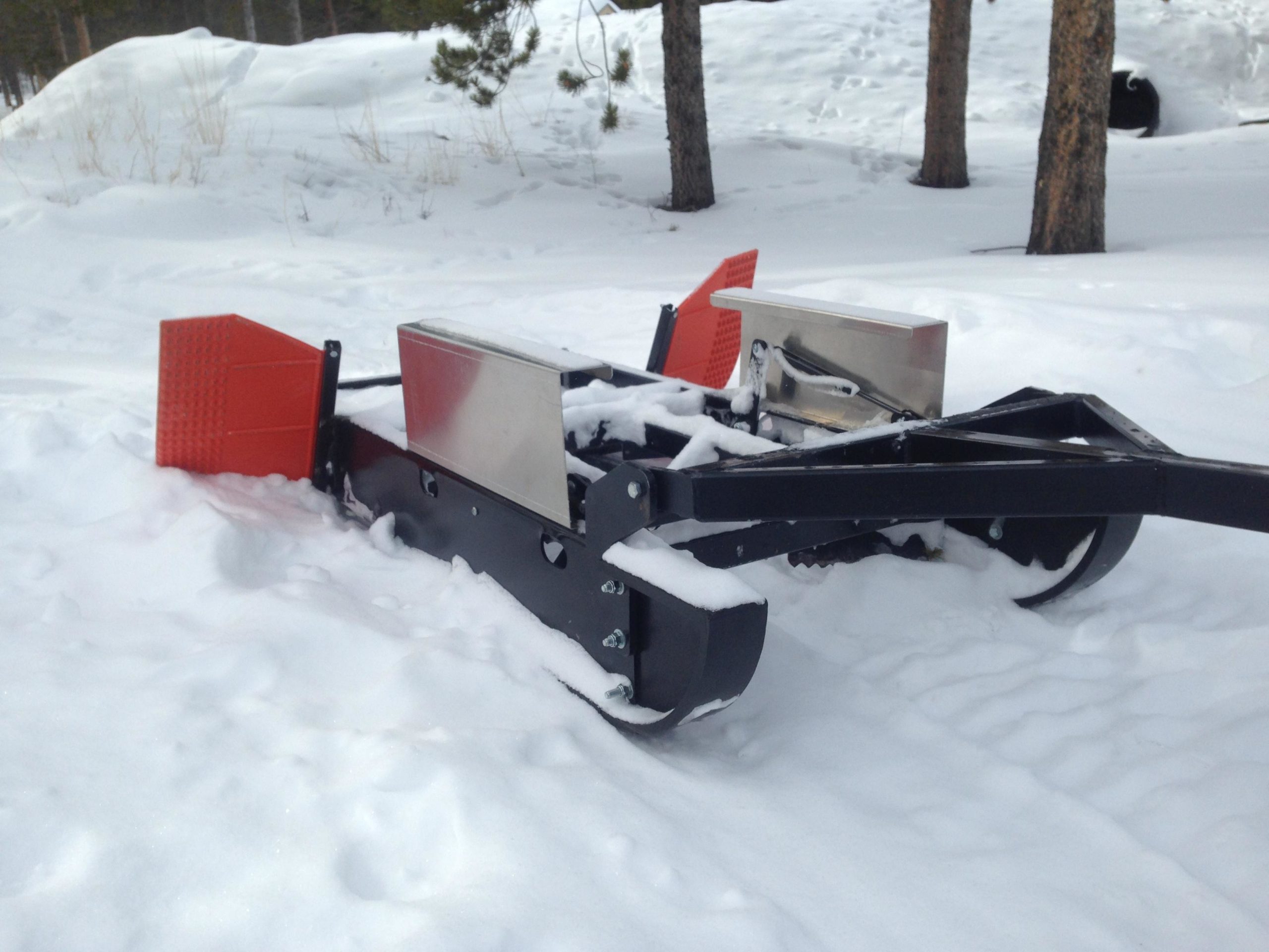 A snow grooming machine partially buried in snow, featuring a black chassis with metal and orange blades, set in a snowy landscape surrounded by trees. Colorado Mountain College Trails mountain bike trail.