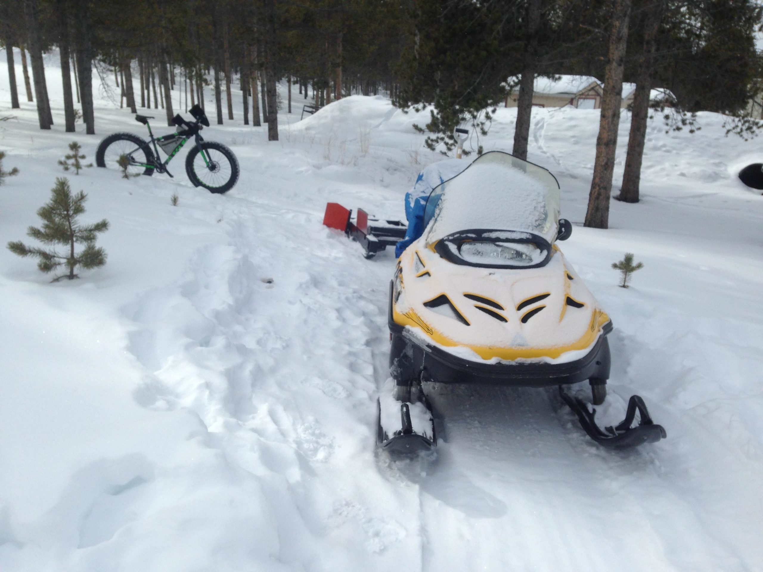 A snow-covered landscape featuring a yellow snowmobile parked on a path through deep snow, with a green fat tire bike leaning against a tree in the background. Pine trees are visible, adding to the winter scenery. Colorado Mountain College Trails mountain bike trail.