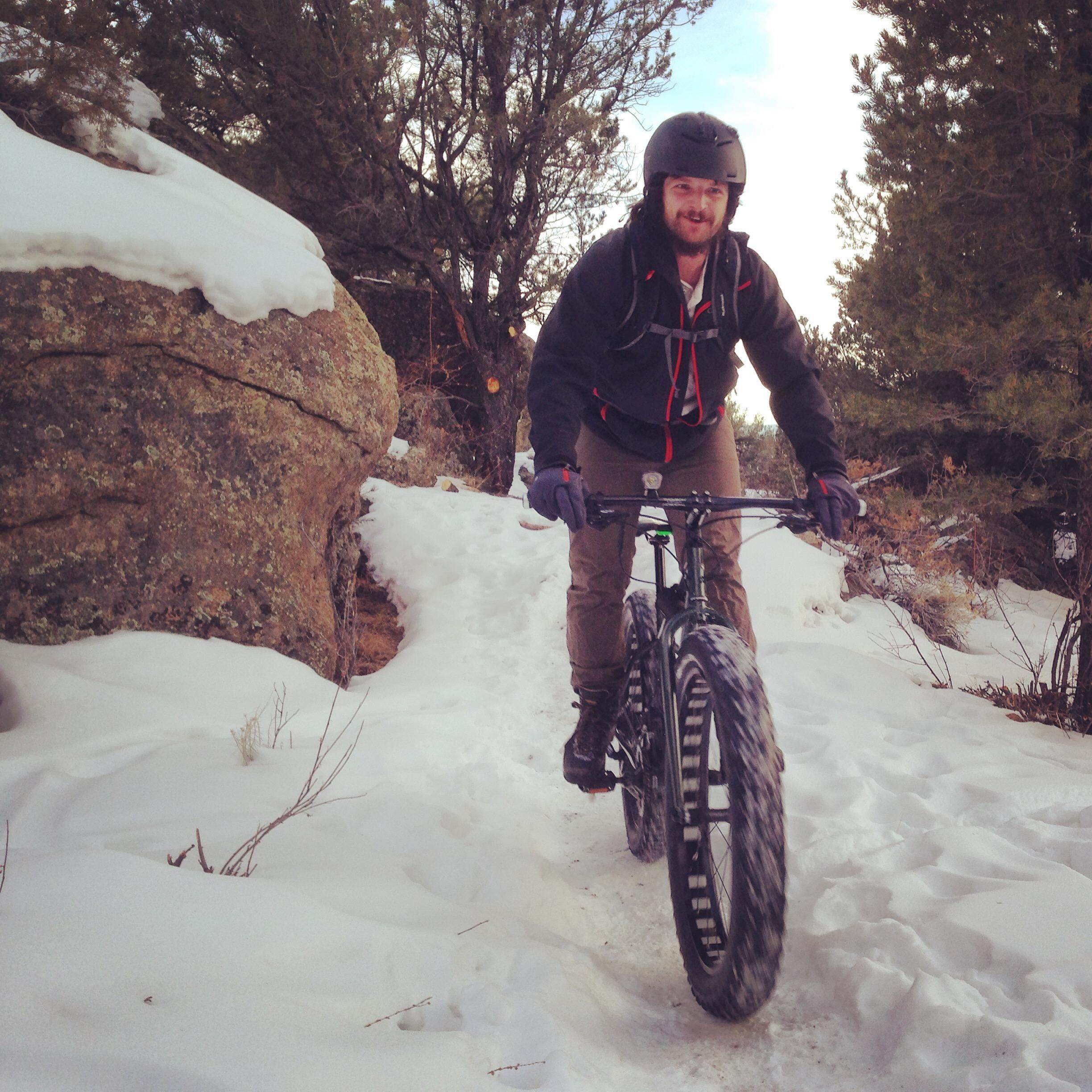 A person riding a fat bike along a snowy trail, surrounded by trees and rocky terrain. The rider is wearing a helmet, gloves, and outdoor clothing, smiling as they navigate the snowy path. Midland Hills Trails mountain bike trail.