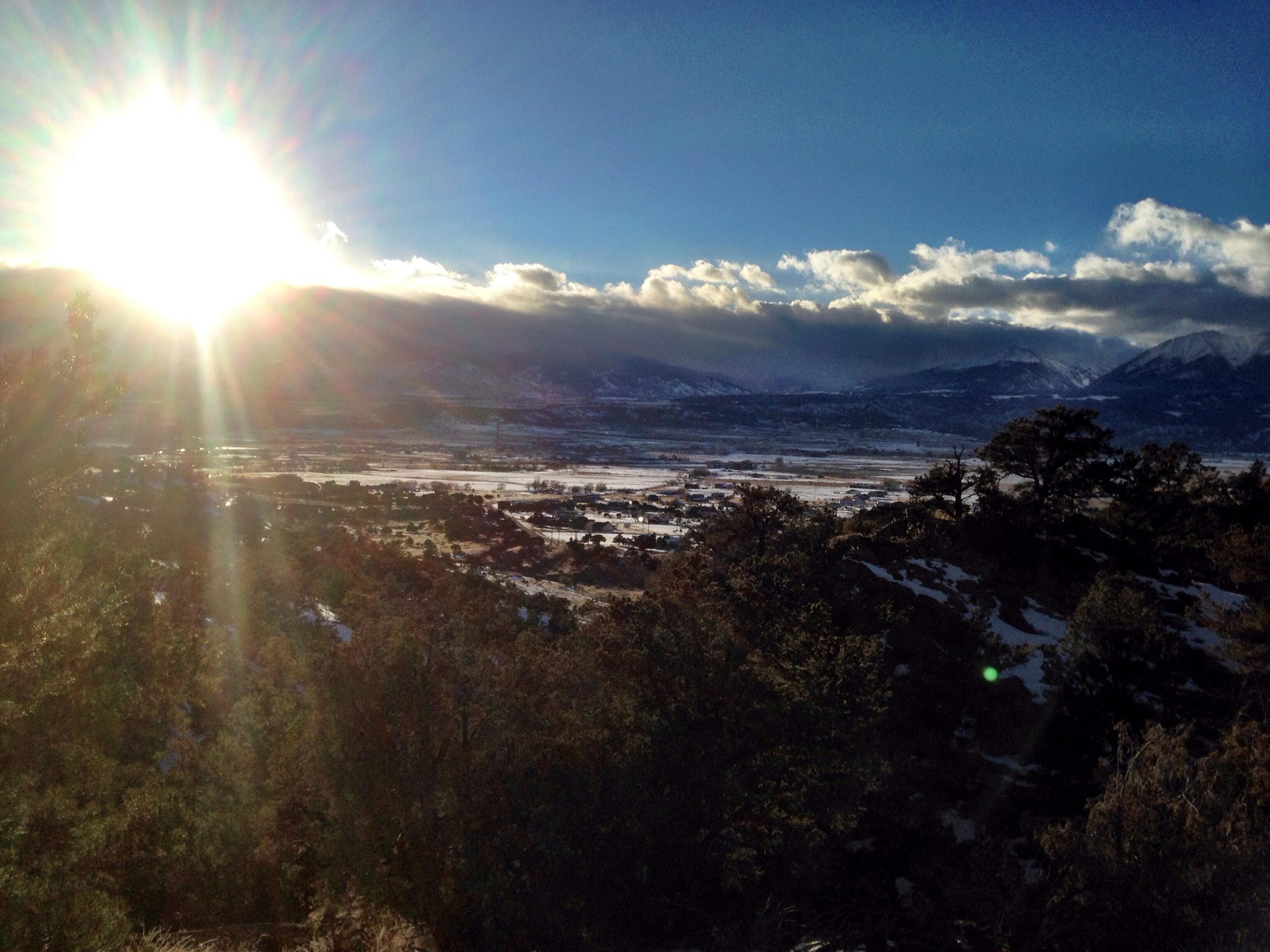 A glowing sun rises over a mountainous landscape, casting light on a valley covered in patches of snow. Trees frame the scene, and clouds are visible in the sky, creating a serene and picturesque view. North Backbone mountain bike trail.