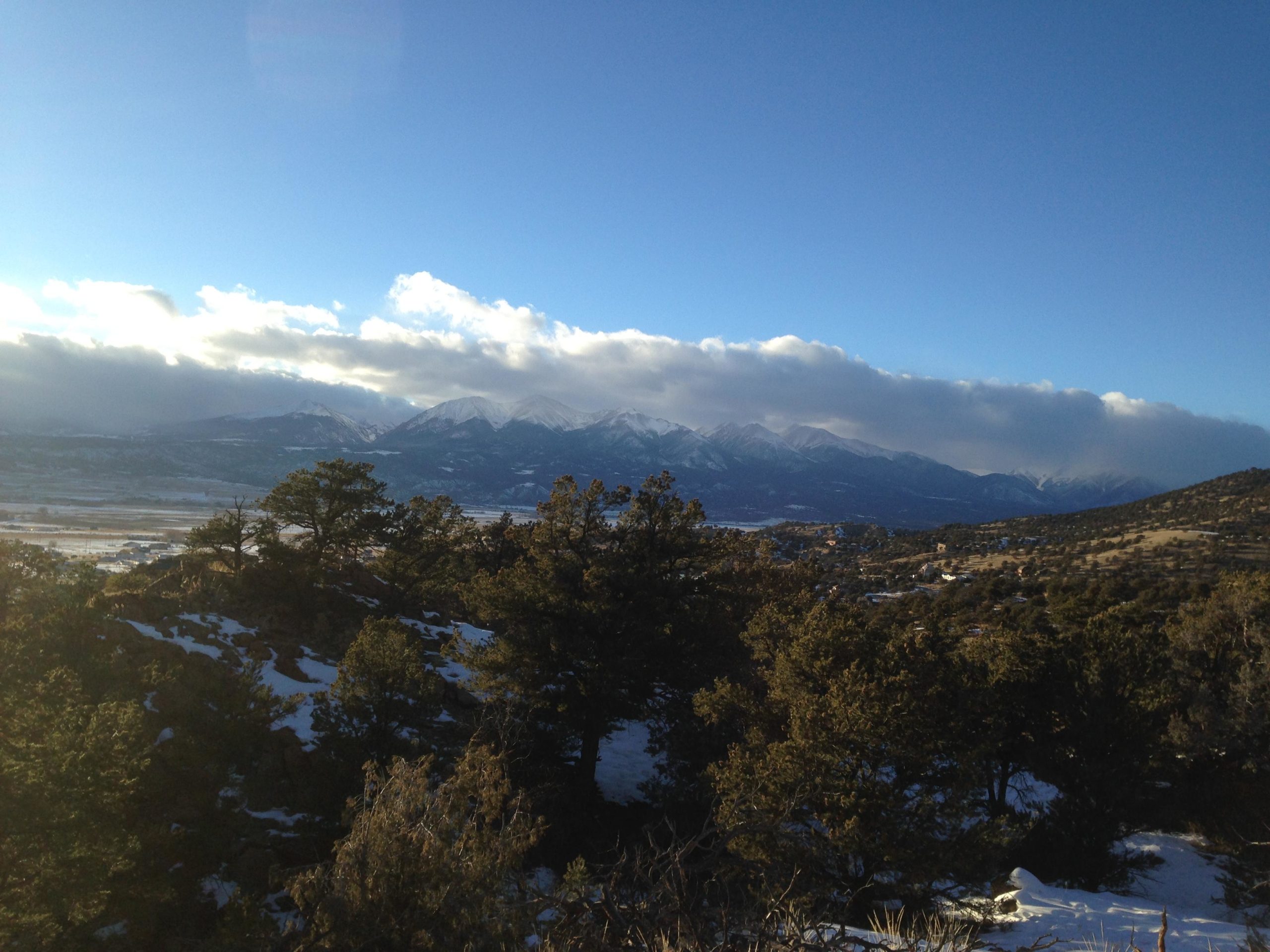 A panoramic view of snow-capped mountains under a partly cloudy sky, with evergreen trees in the foreground and a valley below. The sunlight creates a warm glow on the landscape, highlighting the natural beauty of the scene. North Backbone mountain bike trail.