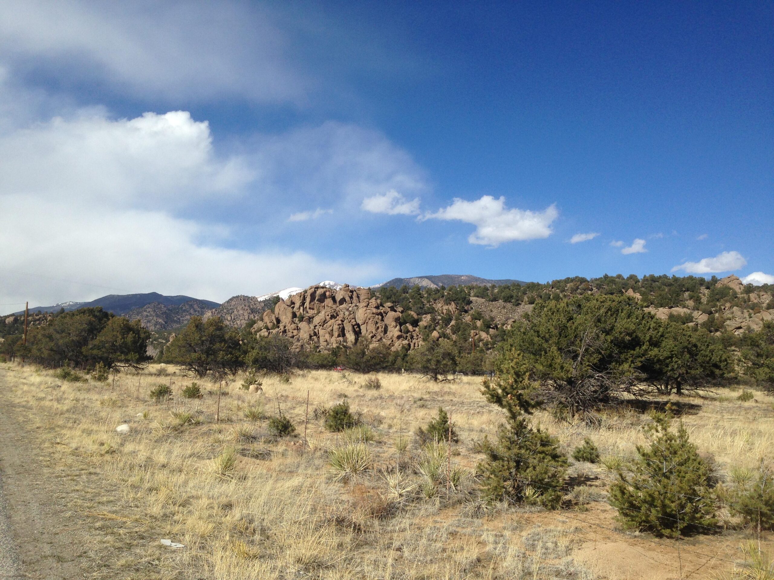 A scenic landscape featuring rocky outcrops and low vegetation under a blue sky with scattered clouds. The foreground includes dry grass and small shrubs, while the background showcases a mountain range, some peaks capped with snow. River Road / #371 mountain bike trail.