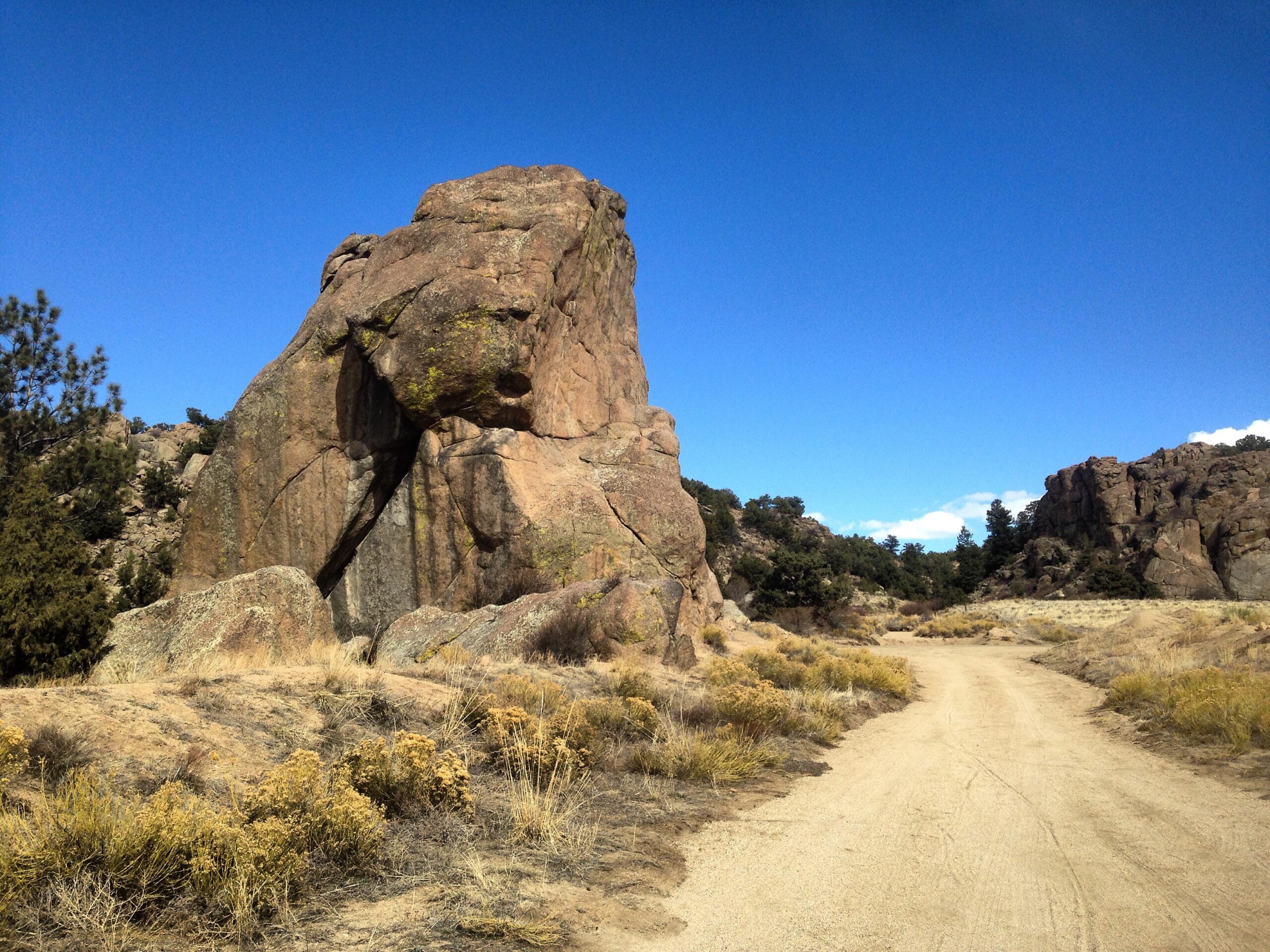 A dirt path winding through a landscape featuring large rock formations, with clear blue skies above and sparse vegetation, including grasses and small shrubs. River Road / #371 mountain bike trail.