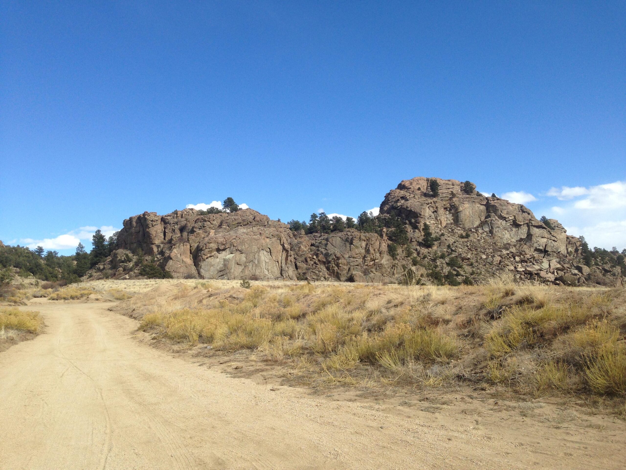 A dirt road leading through a grassy landscape with rocky outcroppings and trees against a clear blue sky. River Road / #371 mountain bike trail.
