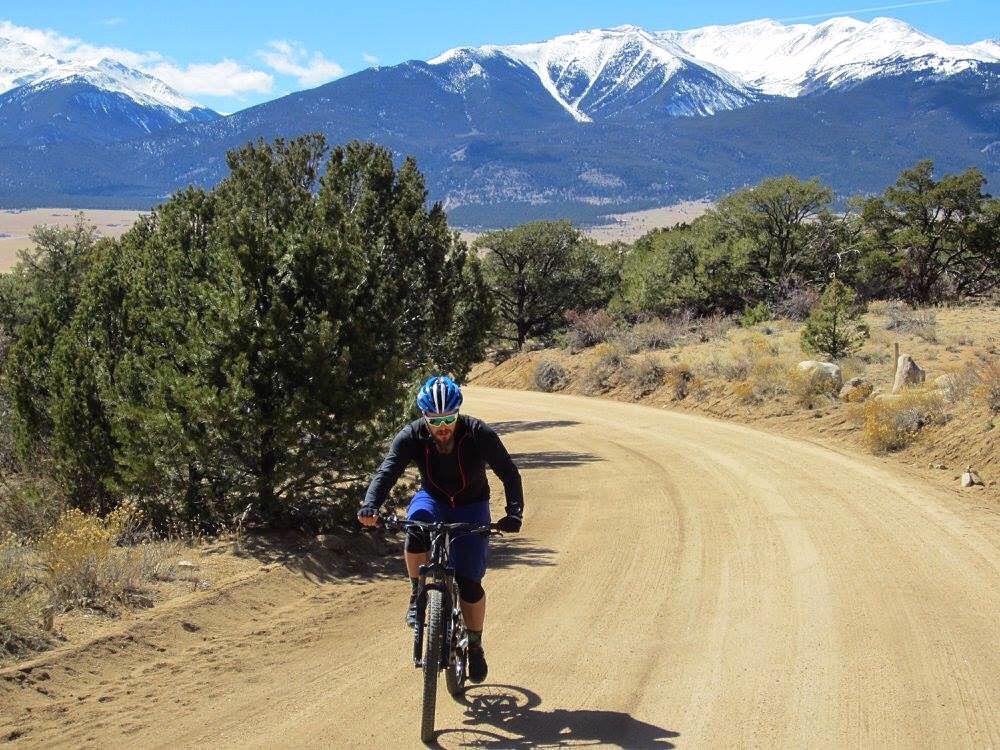 A cyclist riding a mountain bike on a dirt road, surrounded by lush trees with snowy mountains in the background under a clear blue sky. Road #375 mountain bike trail.