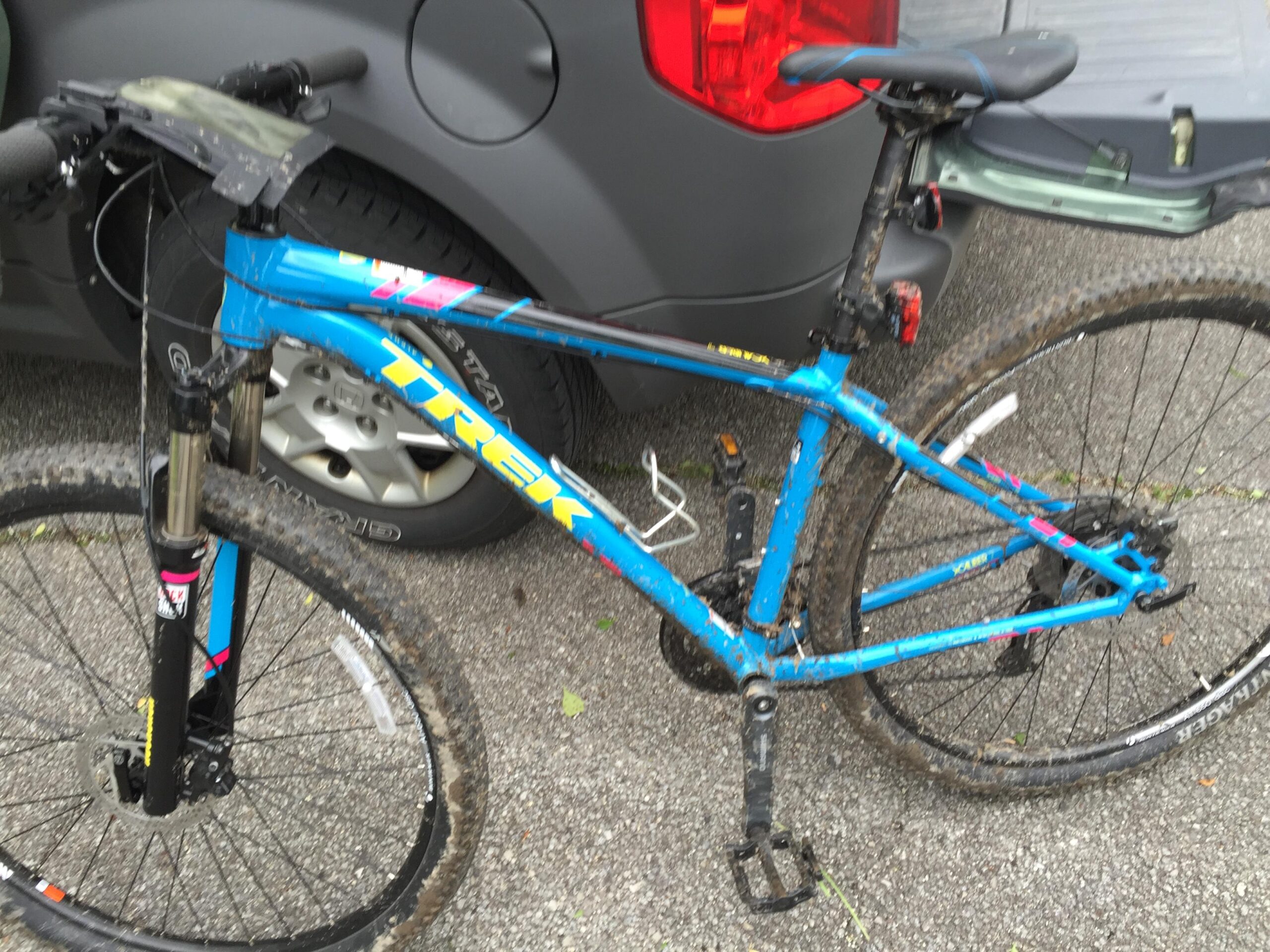 Trek X-Caliber 7: Image of a blue Trek mountain bike with mud on the tires and frame, parked near a vehicle. The bike features front suspension and has a visible gear system. The background includes part of a gray car.