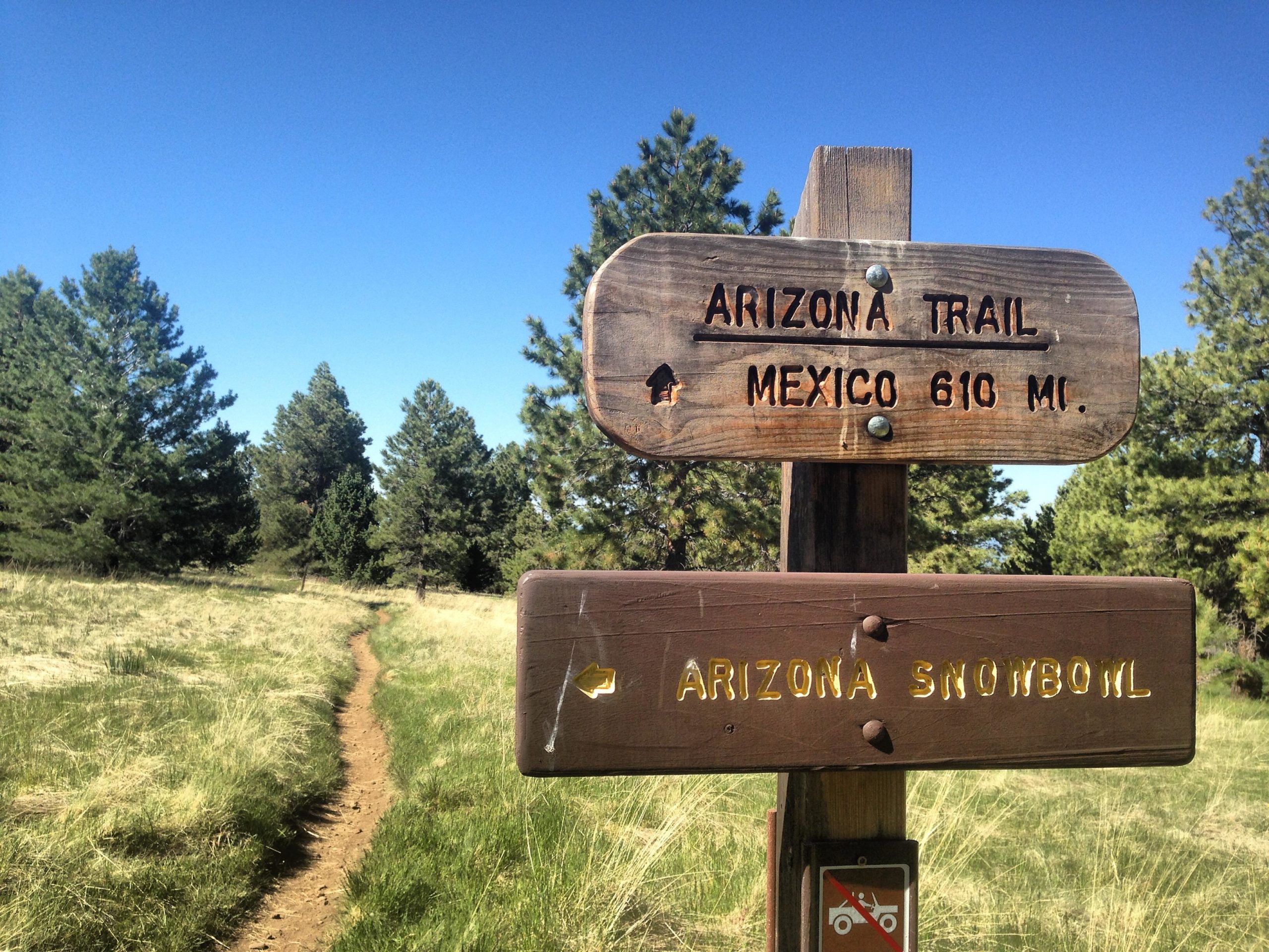 Wooden directional signpost with arrows indicating the Arizona Trail heading toward Mexico, 610 miles away, and a trail to Arizona Snowbowl. Surrounded by grassy fields and evergreen trees under a clear blue sky. Arizona Trail: Flagstaff mountain bike trail.