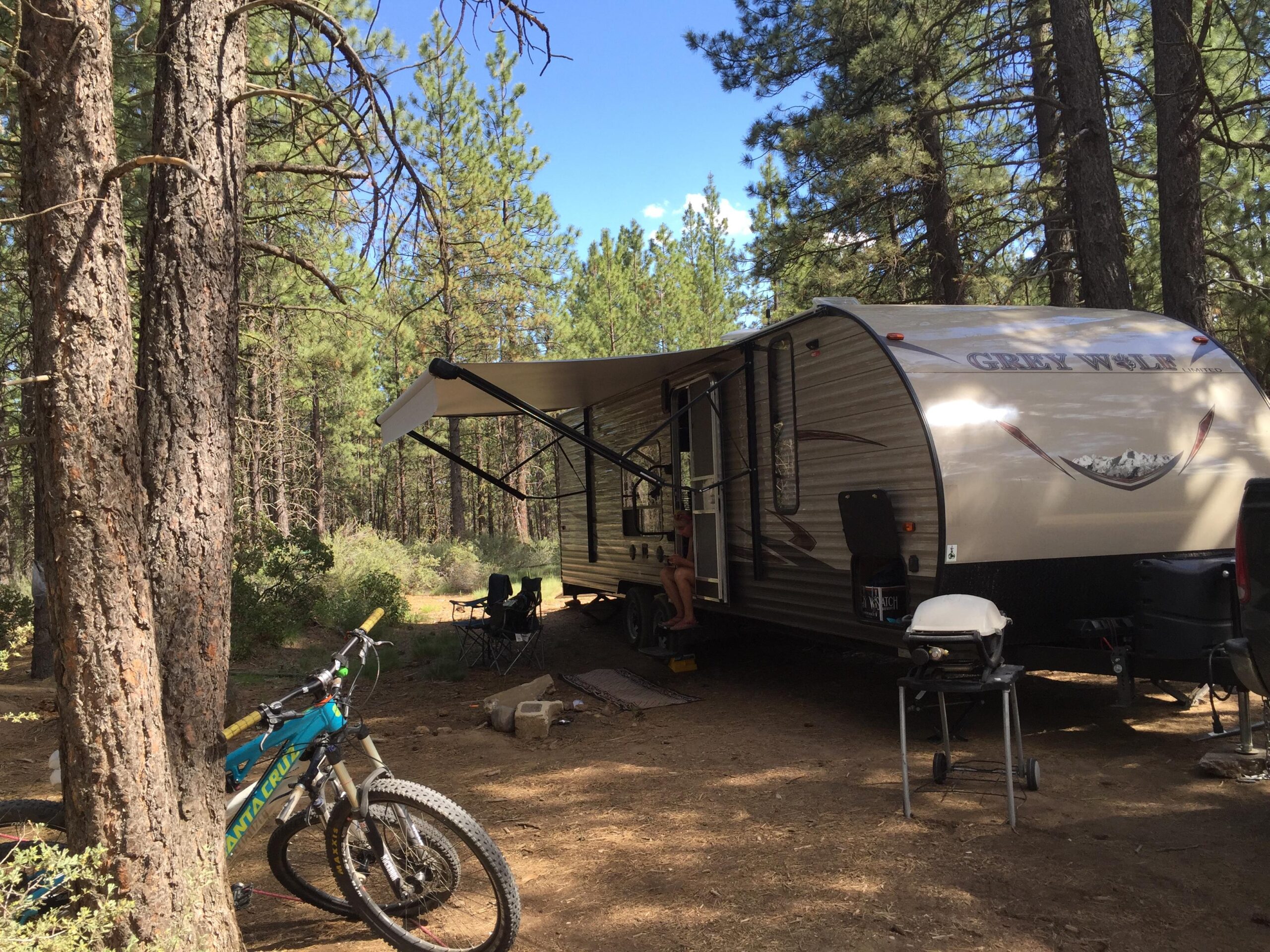 A travel trailer parked in a wooded area, surrounded by tall pine trees. A mountain bike leans against a tree in the foreground, while a grill can be seen on a small table near the trailer. A folding chair is set up nearby, and a person is sitting on the trailer steps, enjoying the outdoor setting. The sky is bright blue with a few scattered clouds. Phil's World mountain bike trail.