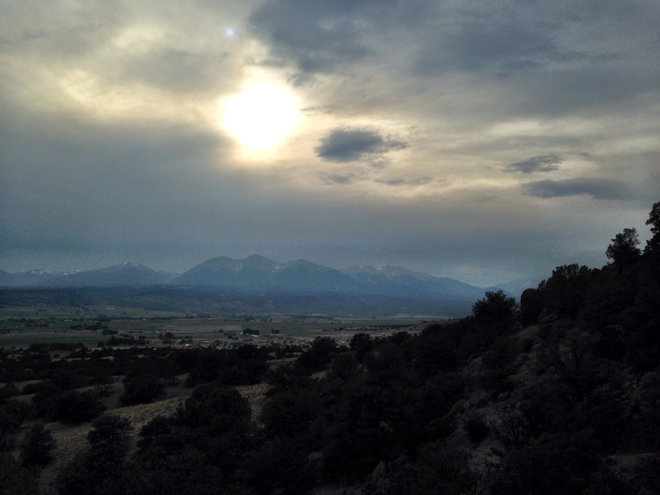 A scenic landscape featuring a range of mountains under a cloudy sky. The sun is partially obscured by clouds, creating a soft, diffused light. The foreground consists of rolling hills covered in sparse vegetation, with patches of trees scattered throughout. The distant mountains have some snow-capped peaks, suggesting a cooler climate. North Backbone mountain bike trail.