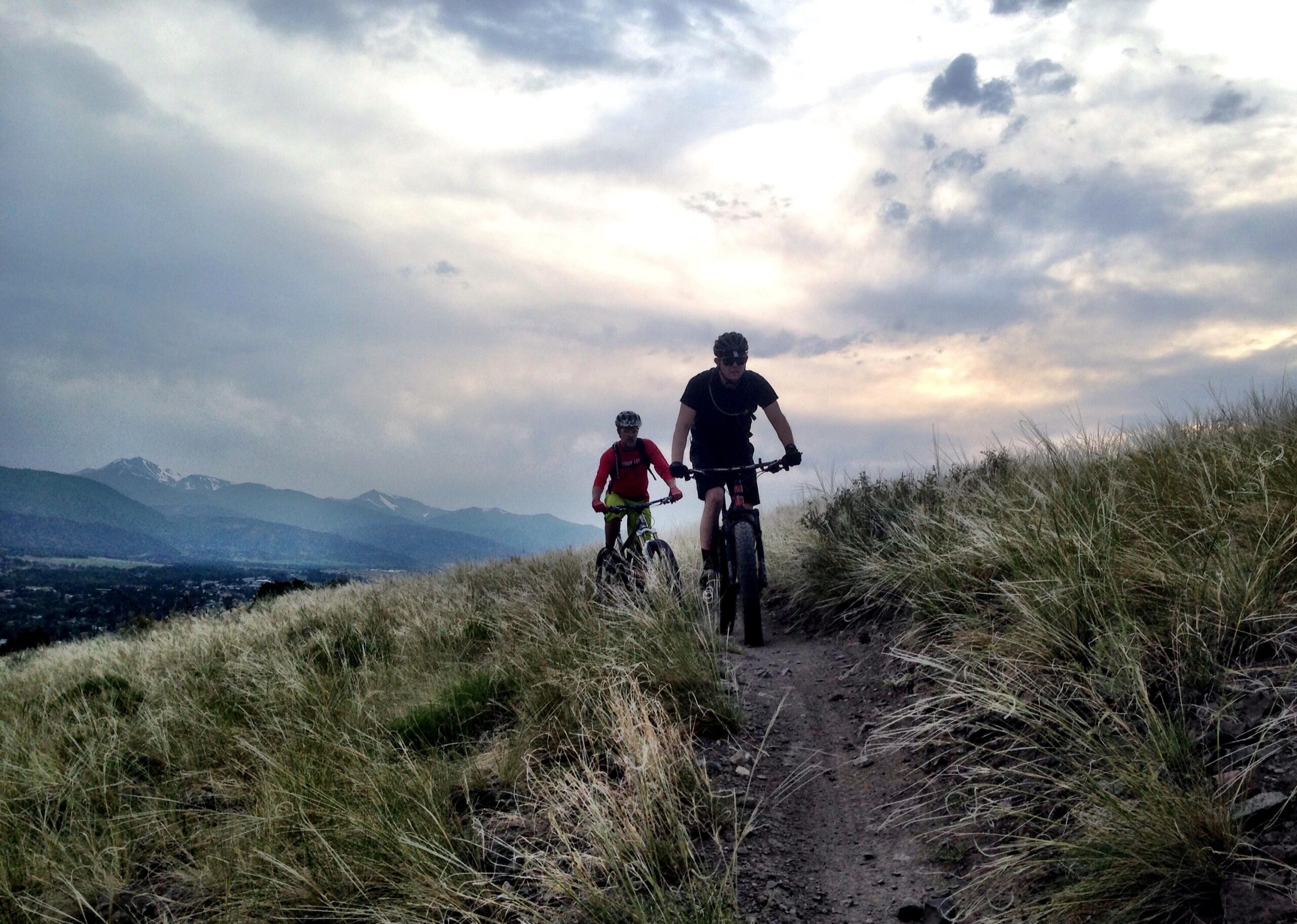 Two mountain bikers riding along a grassy trail with a scenic view of mountains in the background, under a cloudy sky. The rider in front is wearing a black shirt and helmet, while the second rider is dressed in a red jacket and bright green shorts. Arkansas Hills mountain bike trail.
