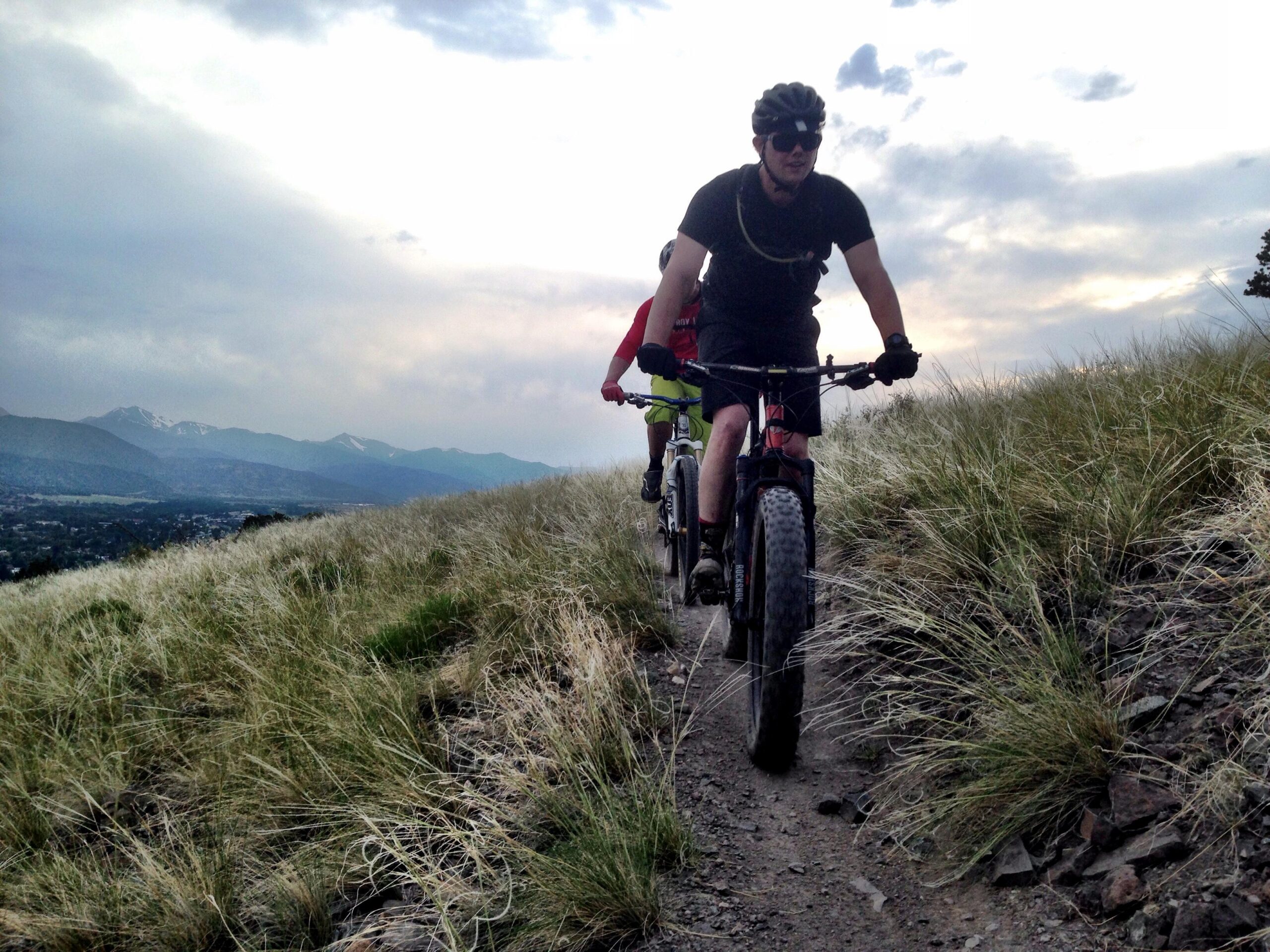 Two mountain bikers riding on a narrow trail through grass and rocky terrain, with a backdrop of mountains and a cloudy sky. The biker in the foreground wears a black outfit and sunglasses, while the second biker, slightly behind, is dressed in a red shirt and green shorts. Arkansas Hills mountain bike trail.