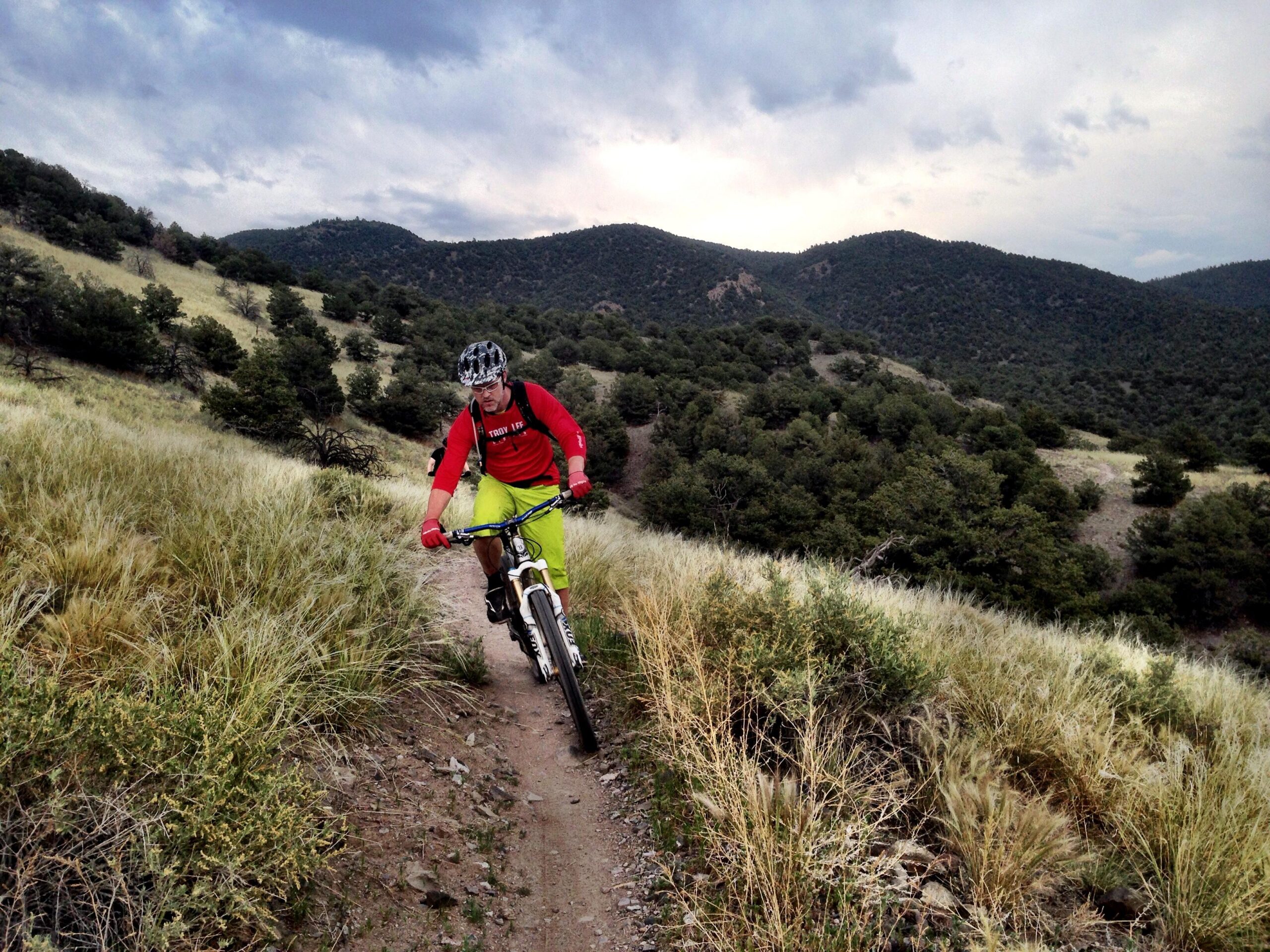 A mountain biker rides along a narrow dirt trail surrounded by tall grass and shrubs, with rolling hills and trees in the background under a cloudy sky. The cyclist is wearing a helmet and bright-colored clothing. Arkansas Hills mountain bike trail.