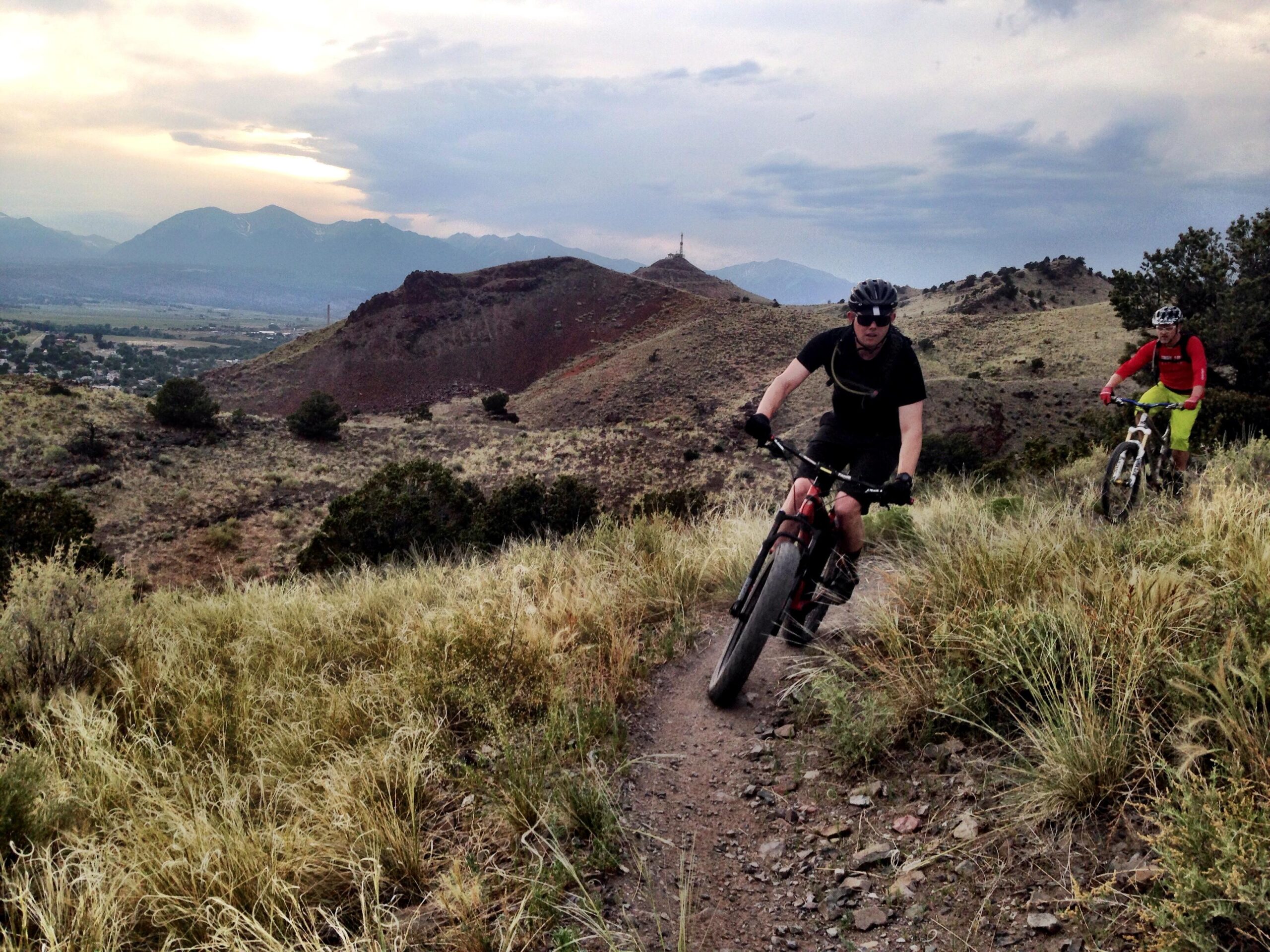 Two mountain bikers navigate a winding dirt trail through grassy hills with distant mountains under a cloudy sky. One rider, dressed in a black shirt and helmet, leans into the turn while the other, wearing a red long-sleeve top and yellow shorts, follows behind. The landscape features sparse vegetation and rocky terrain, creating a sense of adventure in nature. Arkansas Hills mountain bike trail.