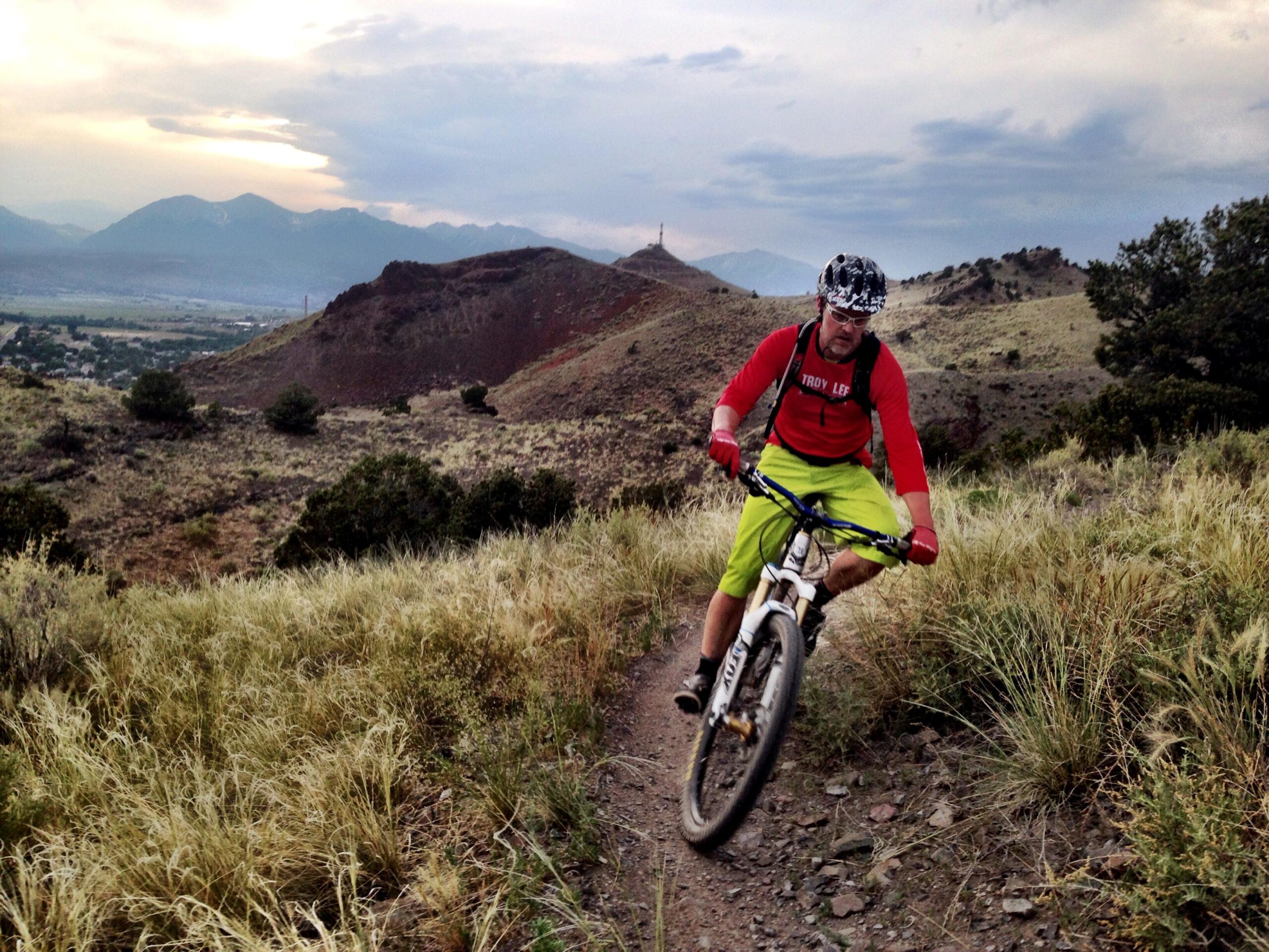 A mountain biker wearing a red long-sleeve shirt and bright green shorts navigates a rugged trail surrounded by tall grass and rolling hills. The background features distant mountains under a cloudy sky, capturing the essence of outdoor adventure. Arkansas Hills mountain bike trail.