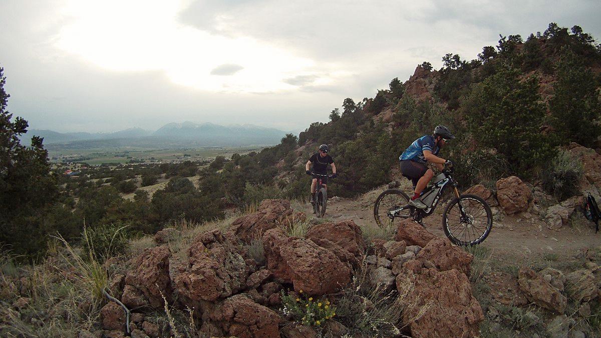 Two mountain bikers navigate a dirt trail surrounded by rocky terrain and greenery, with a backdrop of mountains and an overcast sky. The scene captures the thrill of outdoor cycling amidst nature. North Backbone mountain bike trail.