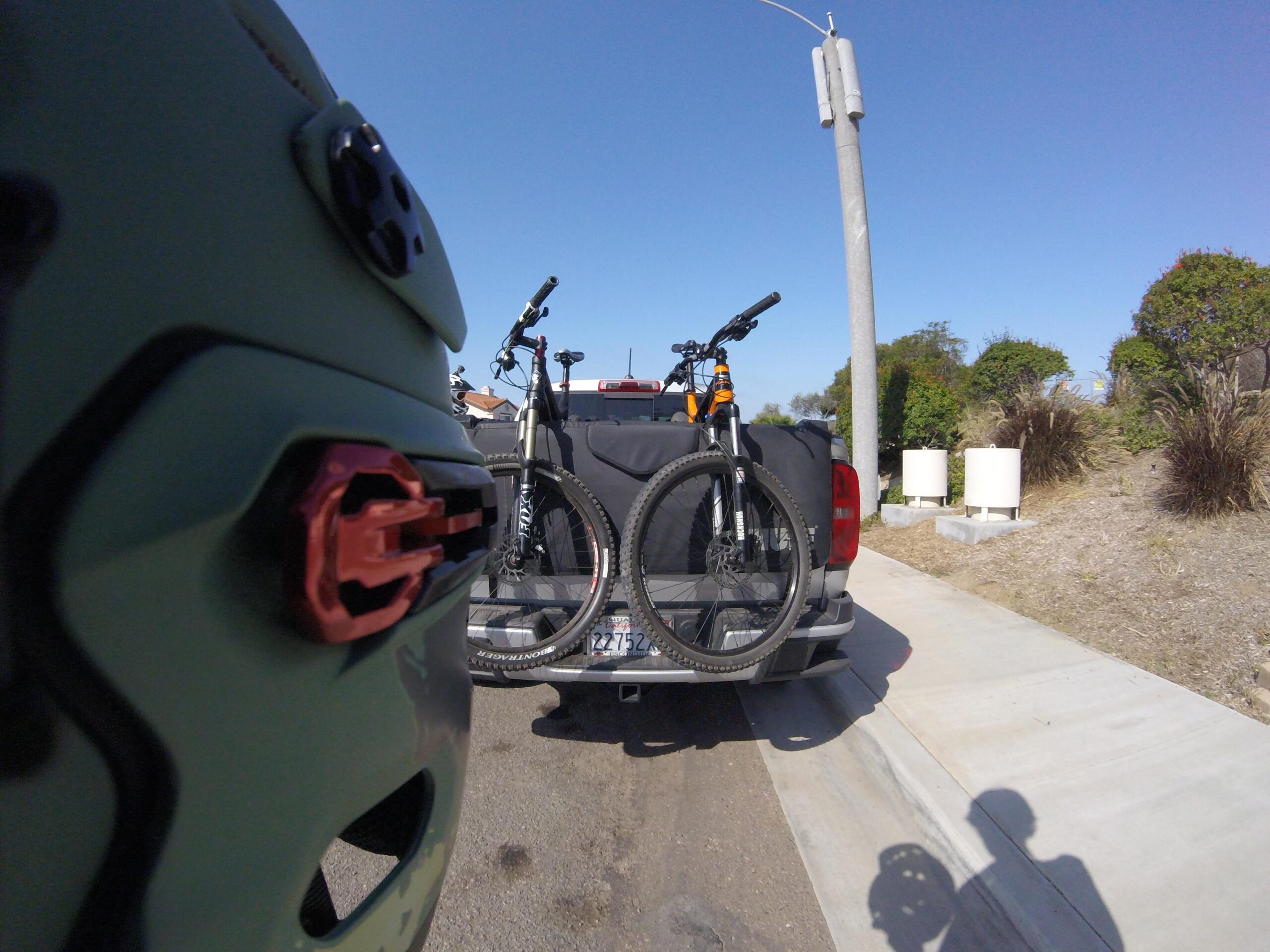 Haro Shift R5: A close-up view of a bicycle mounted on the back of a pickup truck, with a green bike helmet partially visible in the foreground. The scene is set on a sunny day, featuring a clear blue sky and a concrete roadside. Urban greenery is visible in the background.