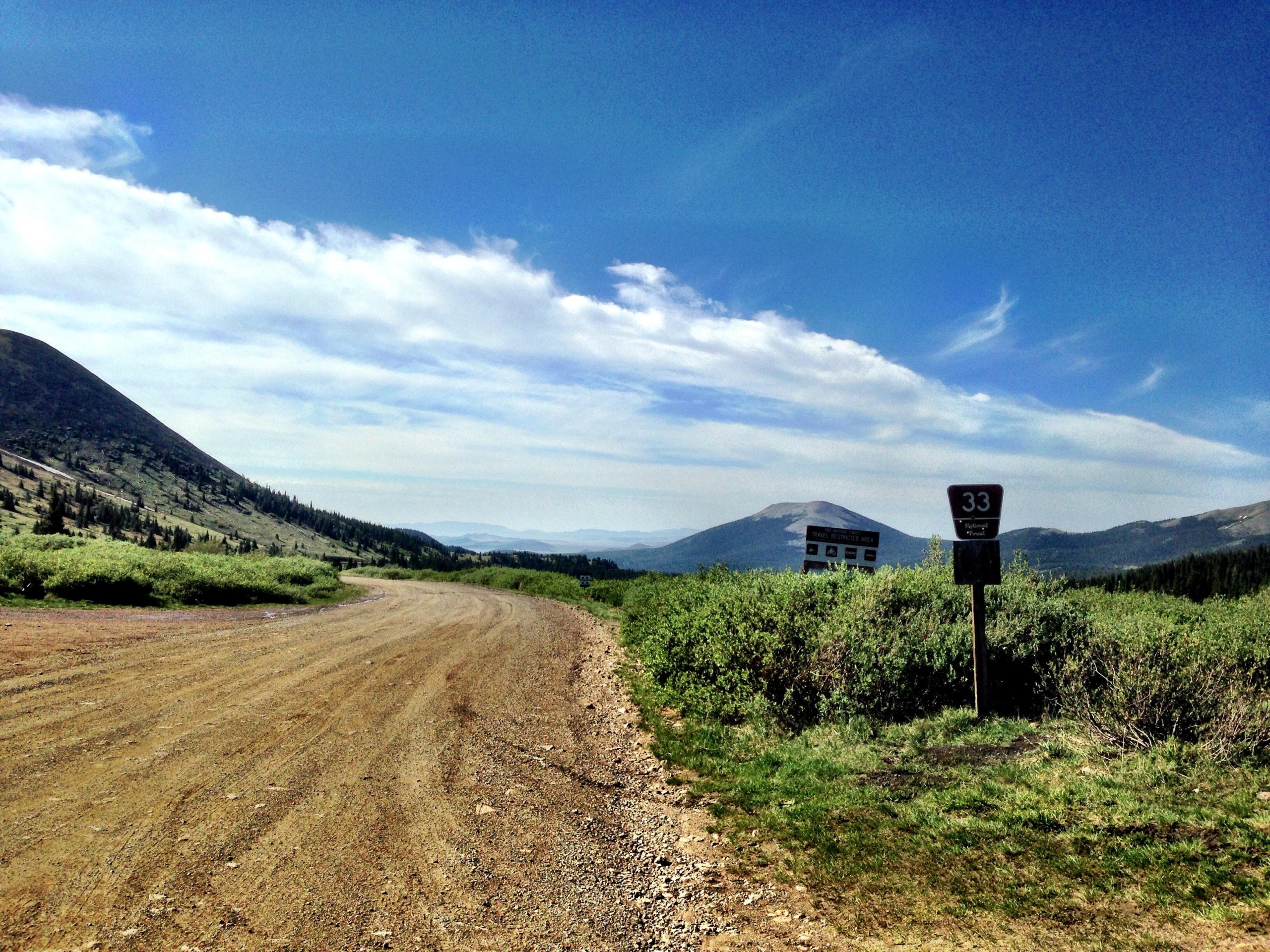 A dirt road winds through a mountainous landscape with lush green vegetation on either side. In the distance, rolling hills are visible under a clear blue sky with wispy clouds. A trail sign labeled "33" stands to the right, indicating a hiking path. The scene conveys a sense of tranquility and natural beauty in an outdoor setting. Gold Dust Trail mountain bike trail.