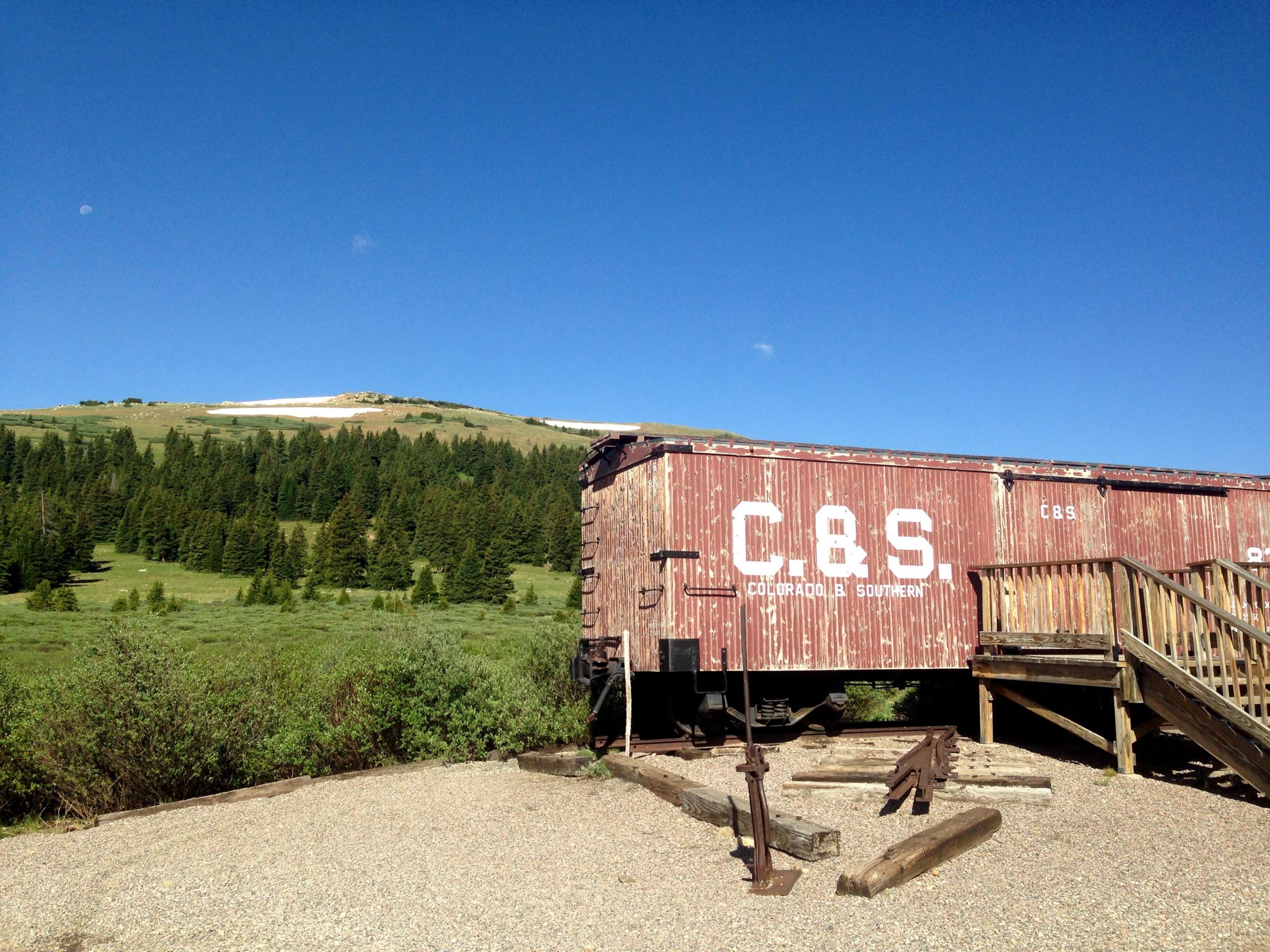 A weathered red train car marked "C&S" is positioned near a wooden staircase, set against a backdrop of rolling green hills and a clear blue sky. A hint of snow is visible on the mountaintops in the distance, and the area is surrounded by lush evergreen trees. Gold Dust Trail mountain bike trail.