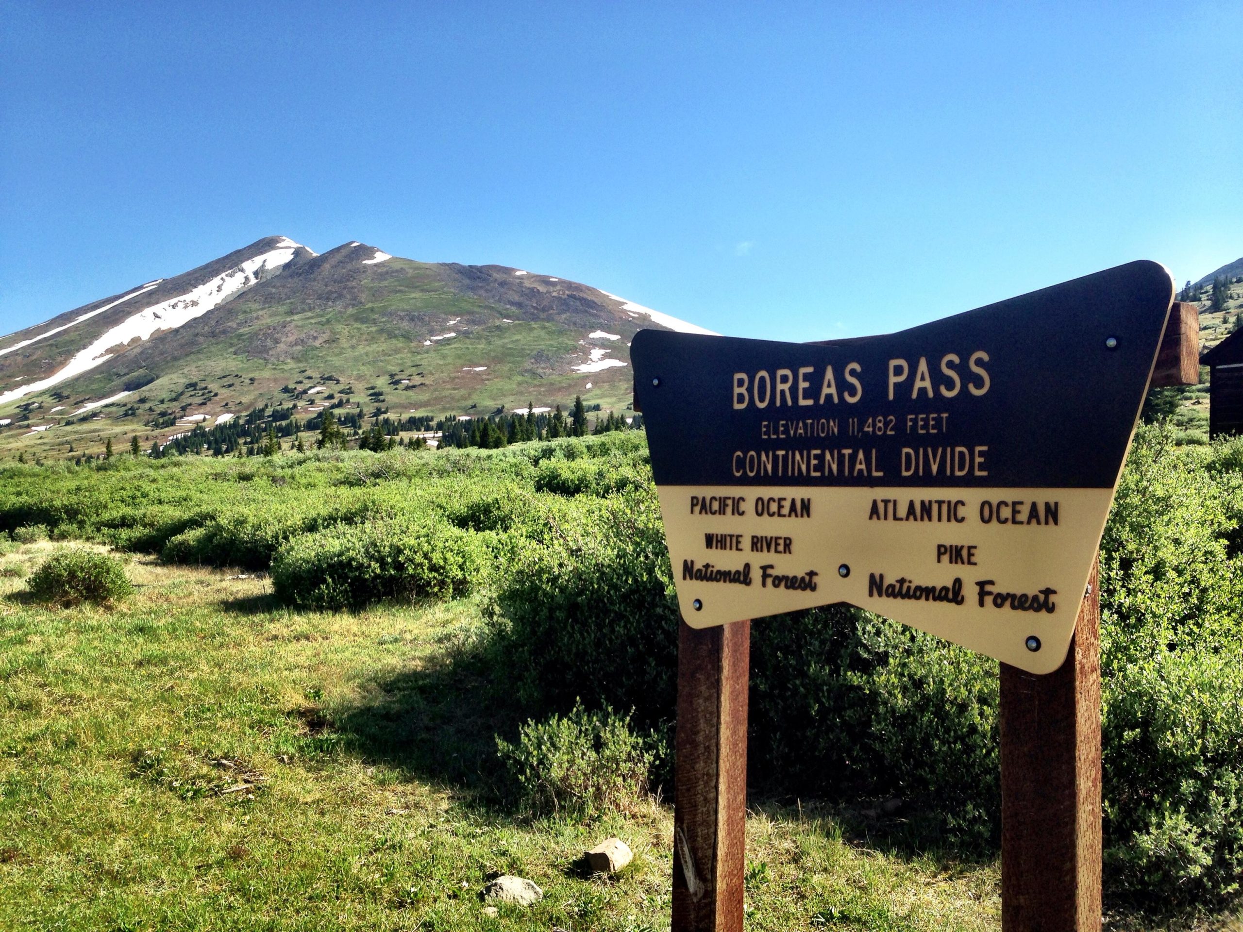 A scenic view of Boreas Pass, featuring a sign indicating the elevation of 11,482 feet and the Continental Divide, with the Pacific Ocean and Atlantic Ocean watersheds noted. The background showcases a snow-capped mountain and lush green meadows under a clear blue sky. Gold Dust Trail mountain bike trail.