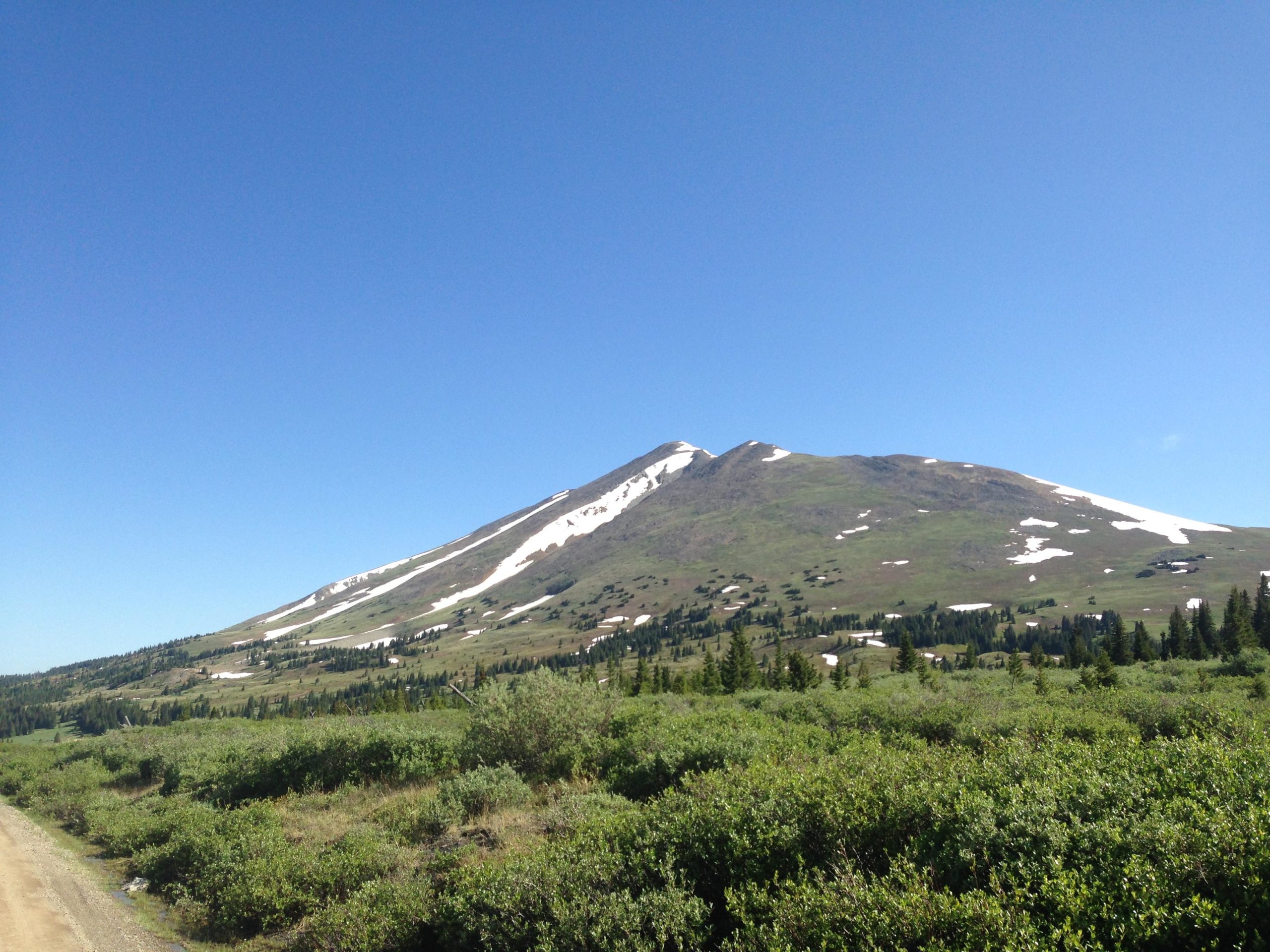 A scenic view of a mountain with a clear blue sky, featuring snow patches on its peak and green vegetation in the foreground. Gold Dust Trail mountain bike trail.