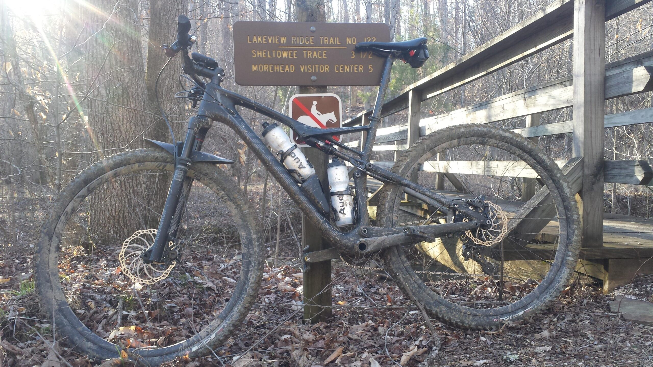 Specialized Epic Expert Carbon World Cup: A mountain bike is leaning against a wooden sign that indicates nearby trails, surrounded by trees in a wooded area. The bike has a muddy frame and two water bottles attached. The sun is shining through the trees, creating a bright atmosphere.