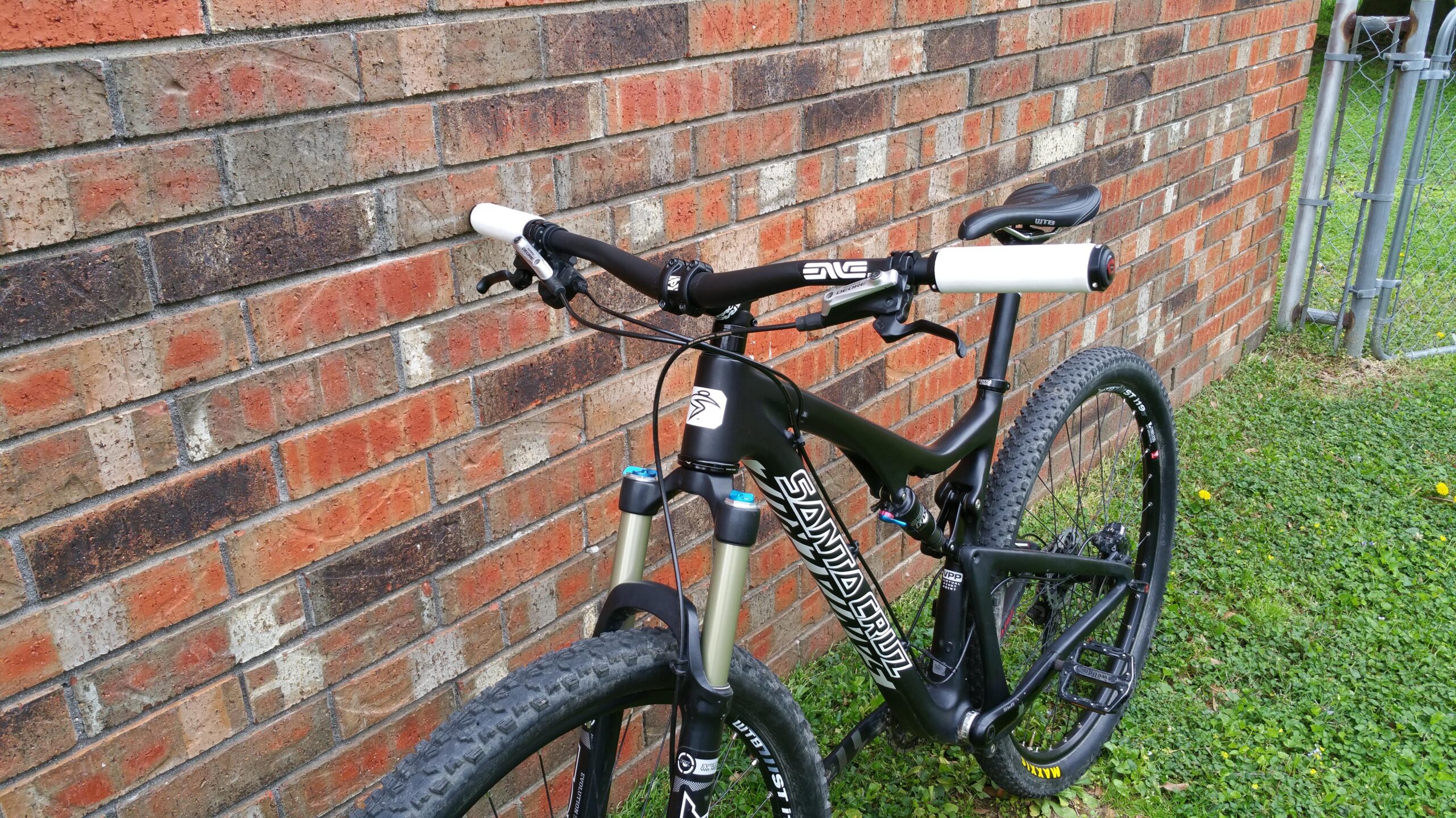Santa Cruz Tallboy Carbon: A black mountain bike leaning against a brick wall. The bike features a sleek frame, wide tires, and white handlebar grips. The front suspension is visible, and the ground is covered in green grass with some yellow flowers.