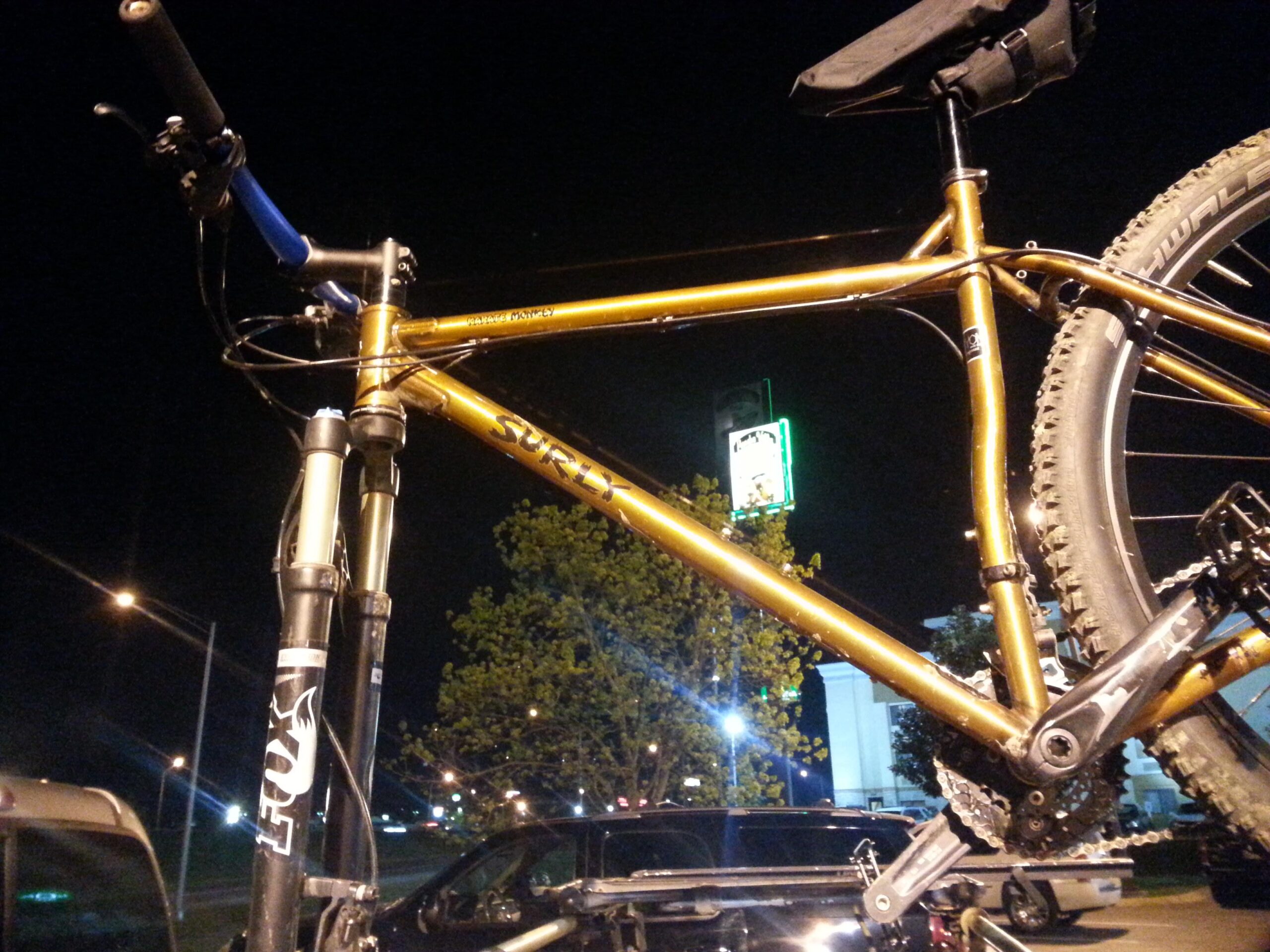 Surly Karate Monkey: Image of a Surly mountain bike with a shiny gold frame, prominently displayed at night. The handlebars with black grips and blue accents are visible, alongside the front fork labeled with a Fox logo. A faintly lit background features a green sign and a glimpse of trees, indicating a nighttime setting.
